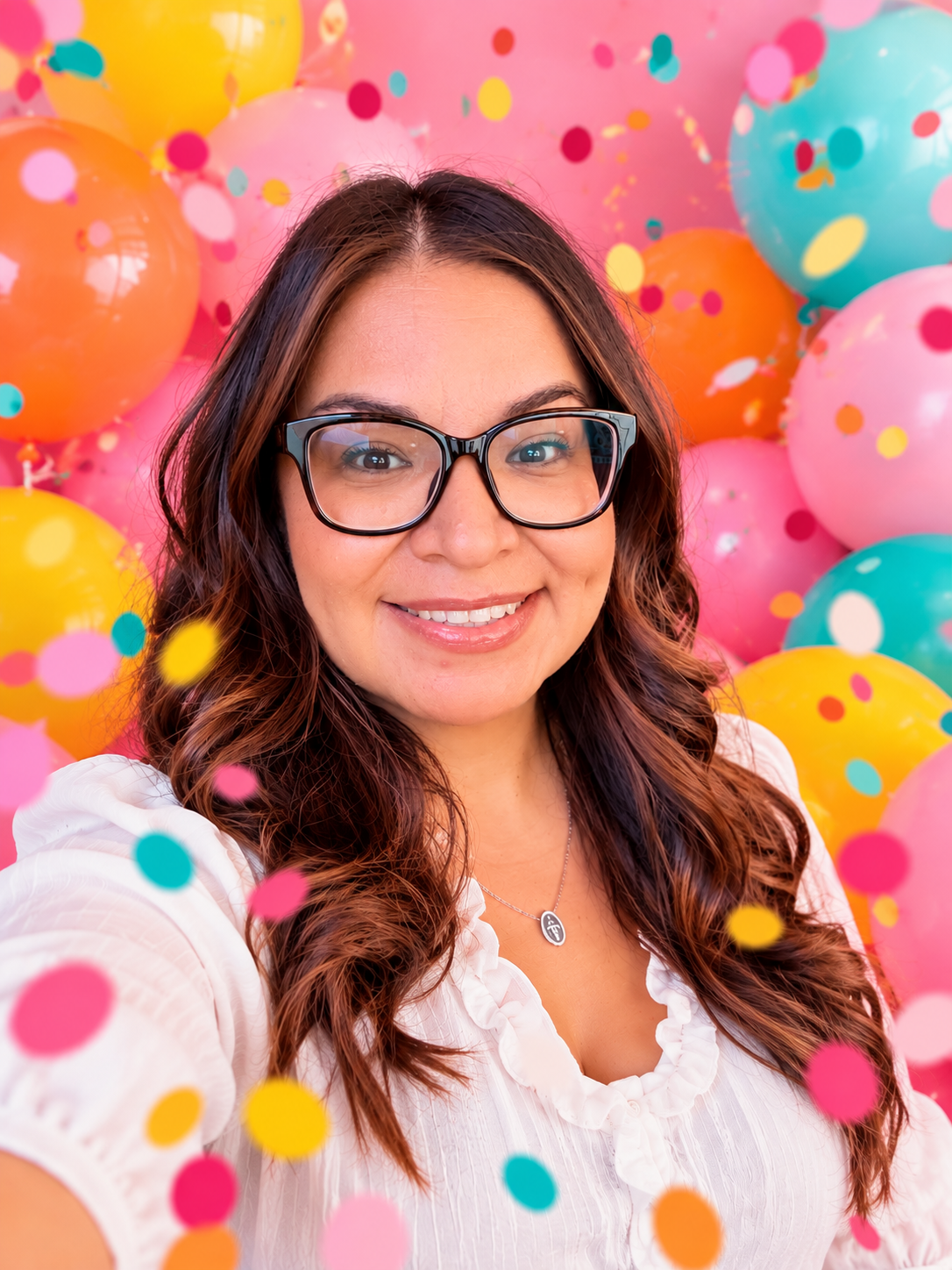 A woman with glasses smiling at the camera, surrounded by colorful balloons and confetti.