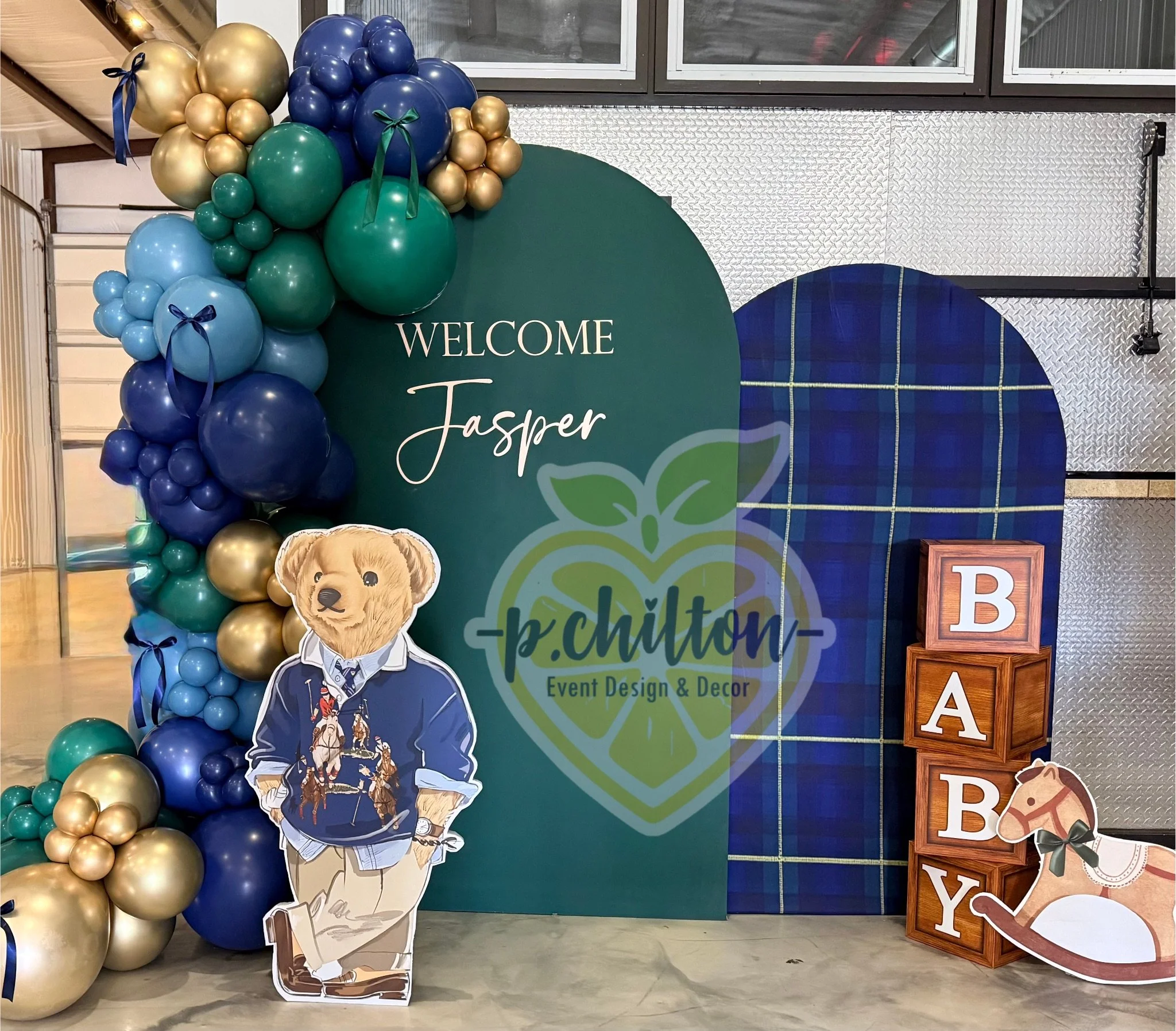 Colorful balloon decoration with shades of gold, green, blue, and navy, along with a cardboard cutout of a stylish dog in a hoodie, at a welcome event for Jasper. There are wooden blocks spelling out 'BEY', a textured metal wall, and a large green and blue sign in the background with event details.