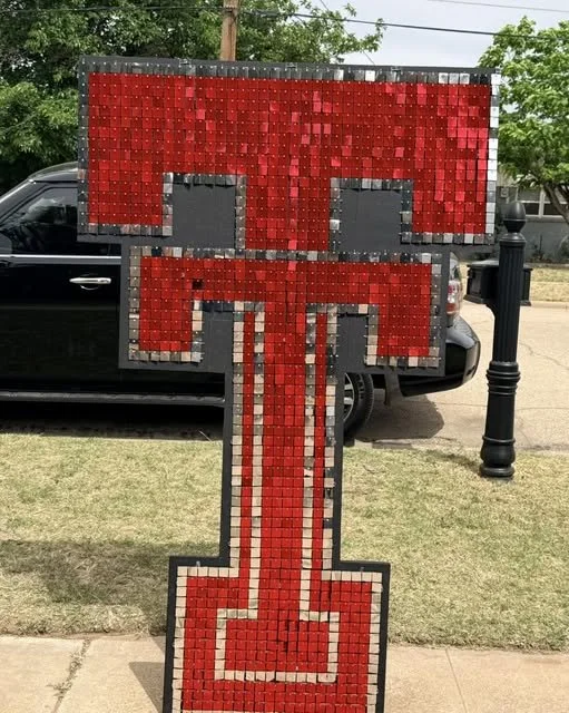 A large sidewalk art piece resembling the letter 'T' with a pixelated design in red, gray, and black, situated outdoors on a grassy area near a street with parked cars.