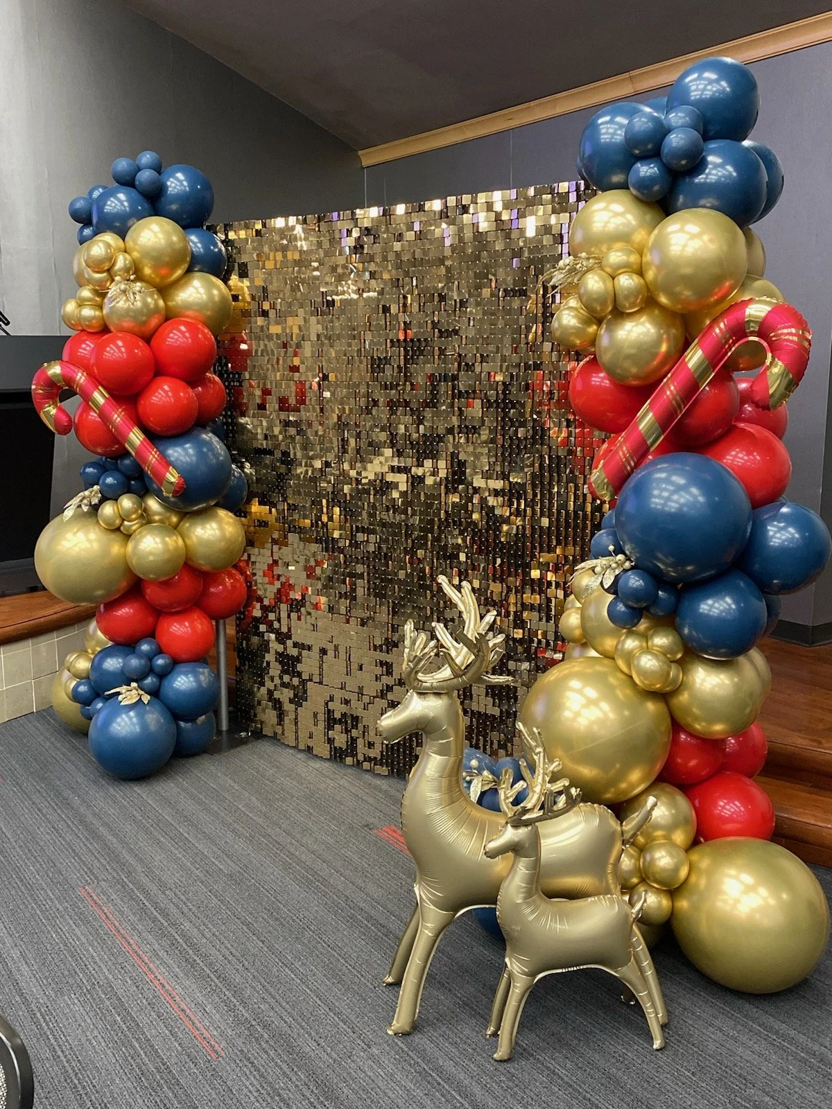 Festive holiday decoration with a gold sequin backdrop, balloons in red, blue, and gold forming an arch, and two gold reindeer balloons in the foreground.