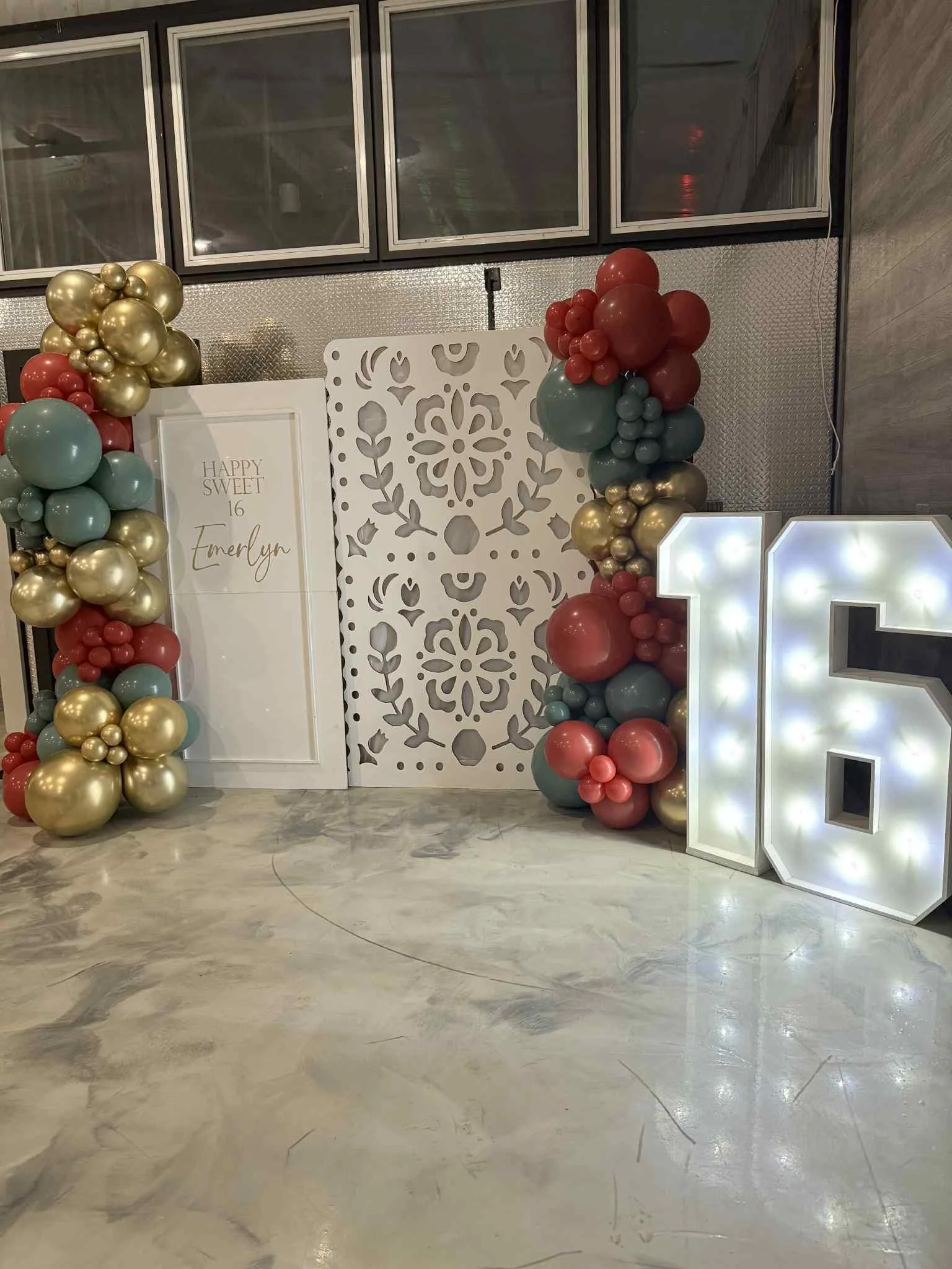 Decorative party backdrop with balloons in gold, red, and teal colors, a white sign with gold lettering saying 'Happy 16 Emily', a white illuminated letter 'L', and a white ornamental panel with cutout floral design, set on a marble floor with a metallic wall and windows in the background.
