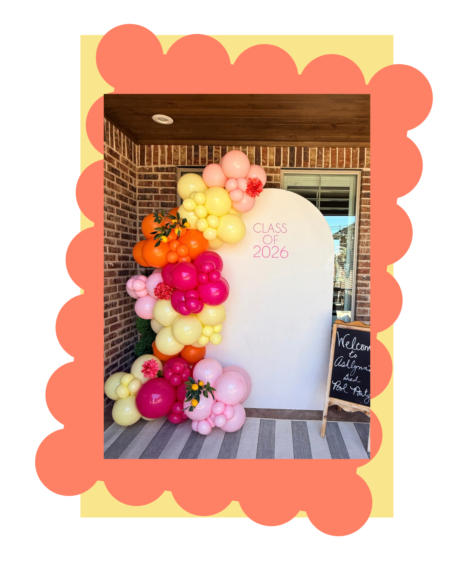 Decorative balloon arch with pink, yellow, orange, and white balloons on a porch with a brick wall, window, and chalkboard sign saying "Welcome to Ashlyn's Grad Pool Party".