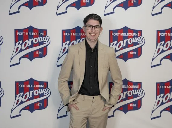 A young man in a beige suit and black shirt standing with hands in pockets, smiling in front of a backdrop with the Port Melbourne Borough logo.