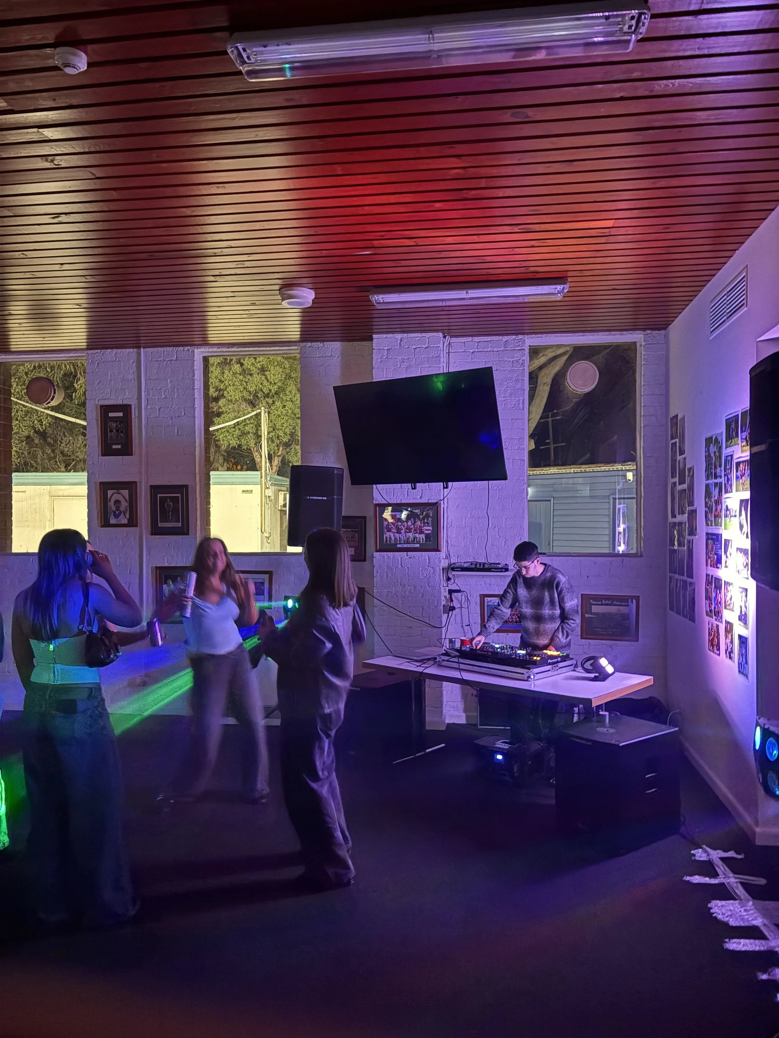People dancing and DJ playing music in a dimly-lit indoor venue with posters on the wall and windows showing outside scenery.