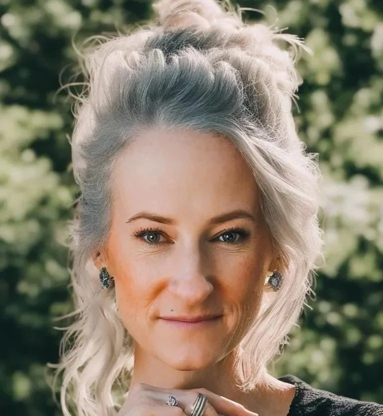 Close-up of a woman with blonde, wavy hair pulled back, wearing earrings, outdoors in a natural setting.
