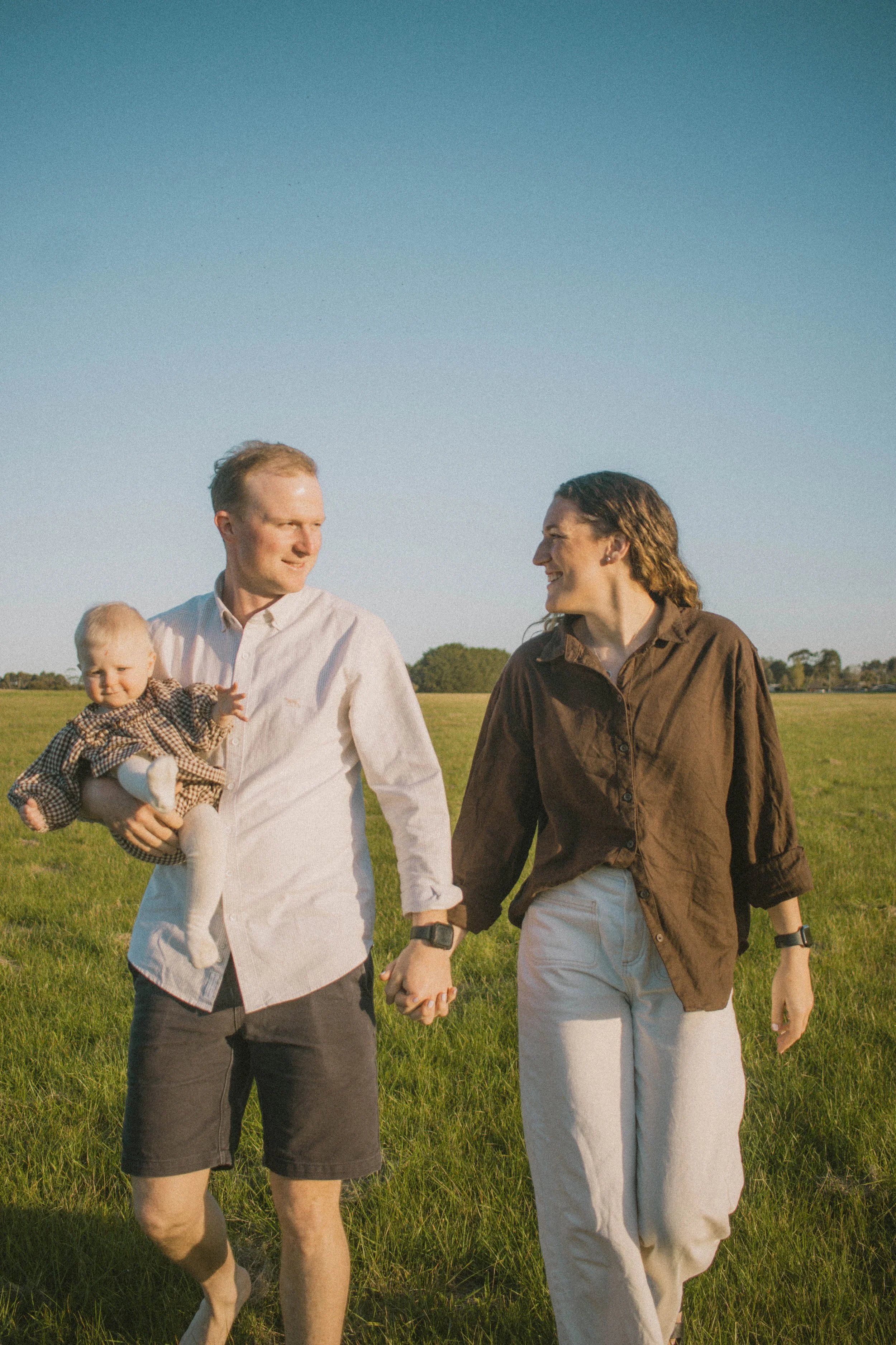 A man wearing a white shirt holding a toddler walking hand in hand with a woman in a brown shirt in a grassy field under a clear blue sky.