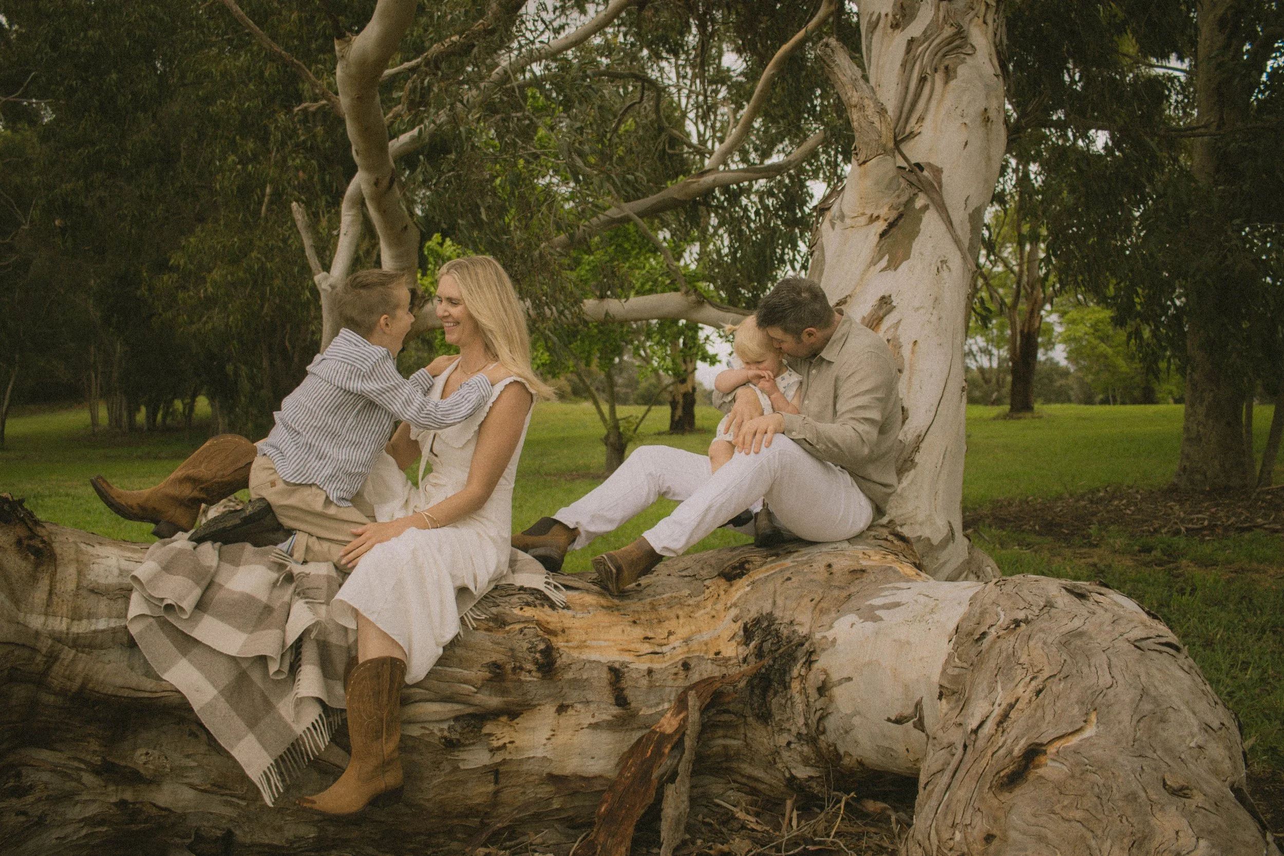 A family of four sitting on a large fallen tree trunk in a park, hugging and smiling at each other, with green trees and grass in the background.