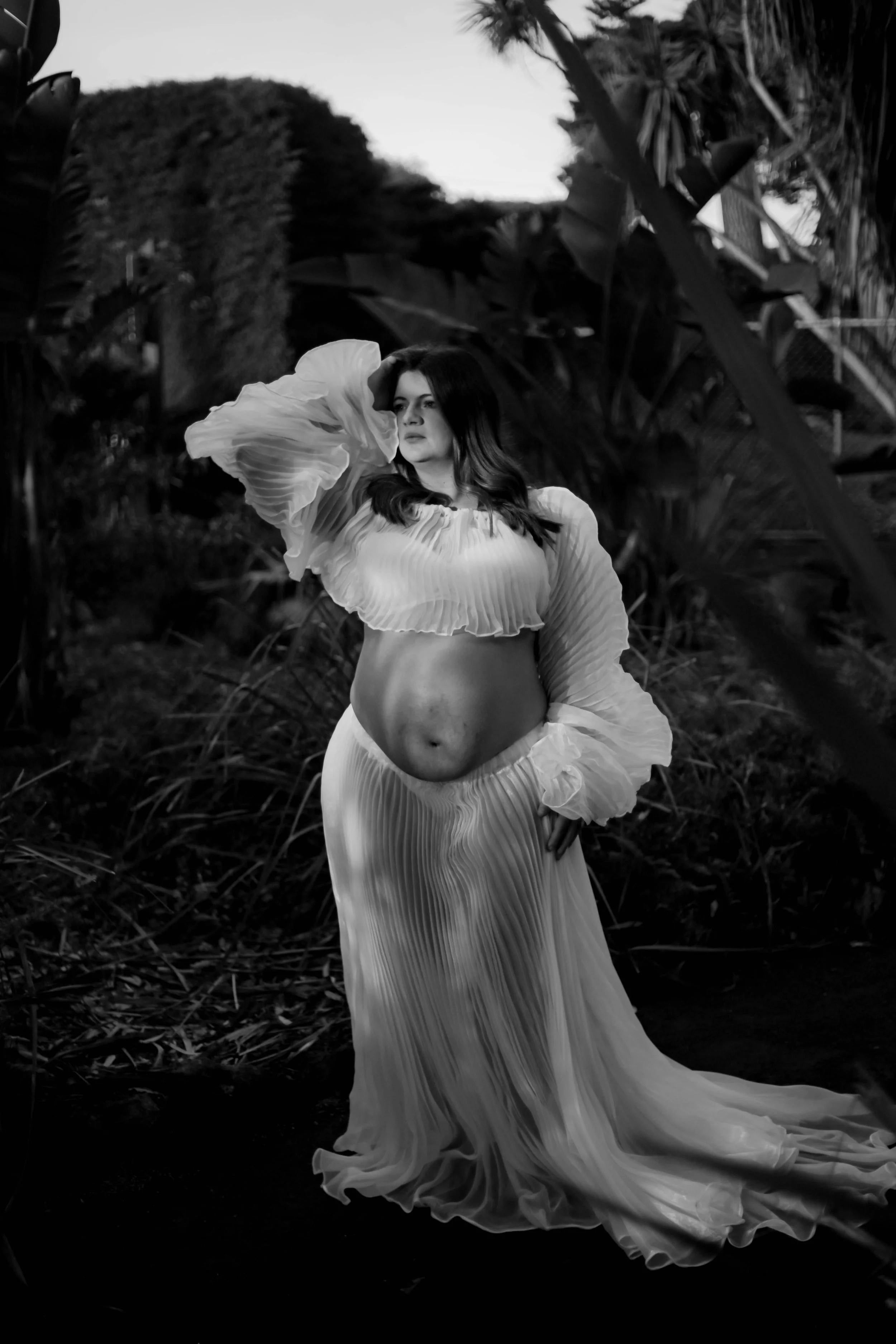 A woman in a flowy, pleated dress stands outdoors amidst large foliage, with her hand resting on her head and looking into the distance.