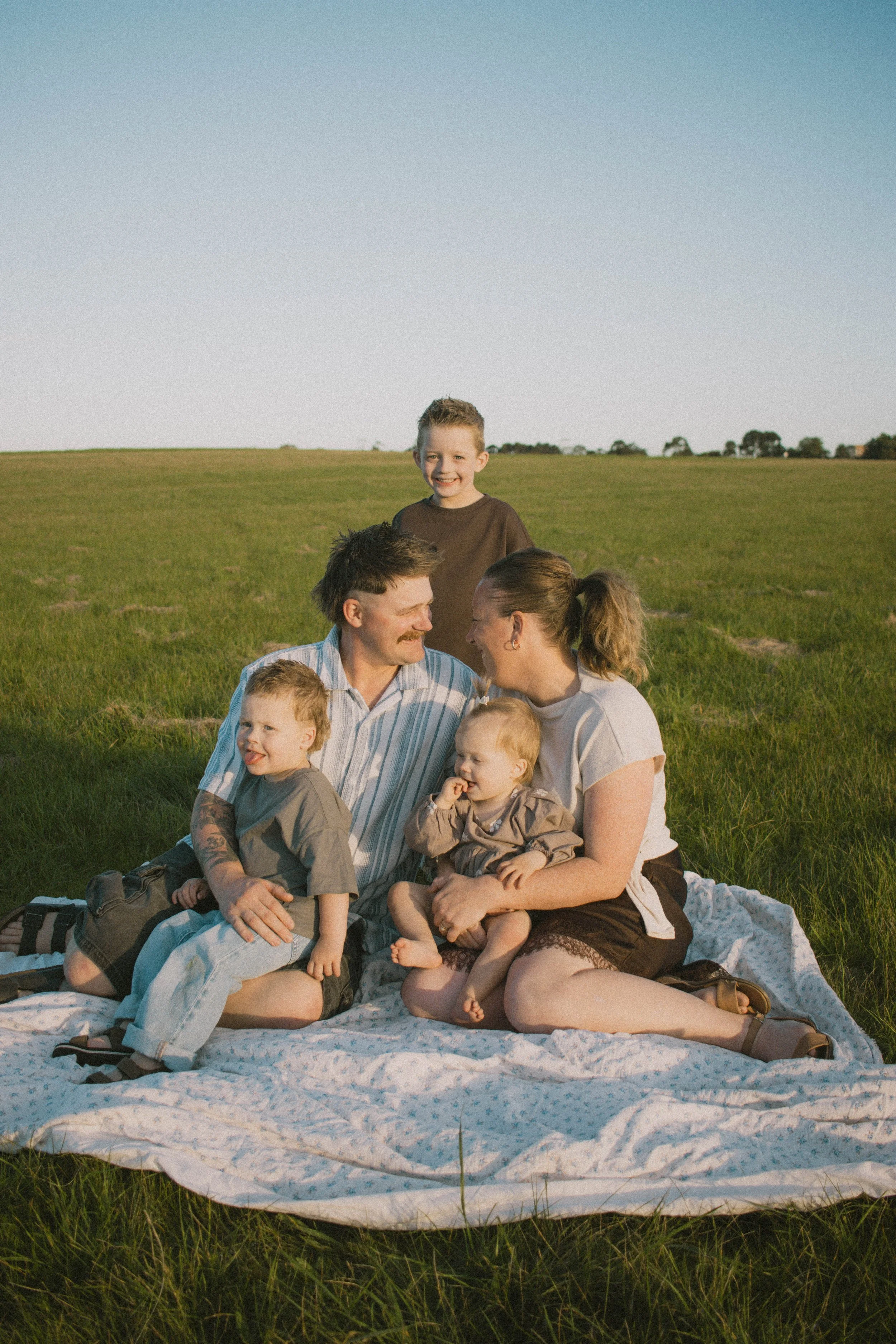 A family of six sitting on a white blanket in a grassy field with a clear sky, smiling and enjoying each other's company during a sunny day.