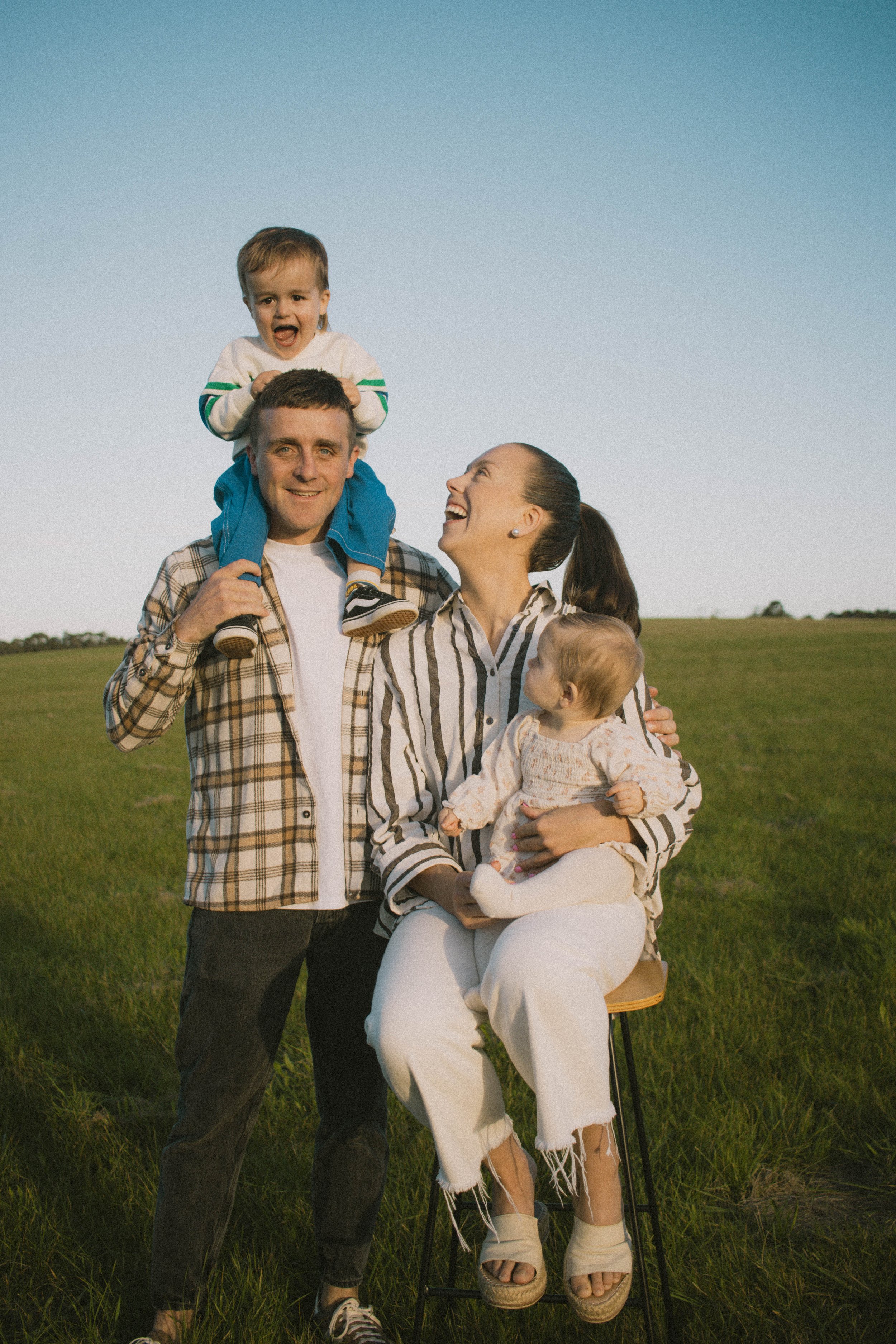 Family of four outdoors in a field with a clear sky, two adults and two children, one child on an adult's shoulders, all smiling and enjoying the moment.