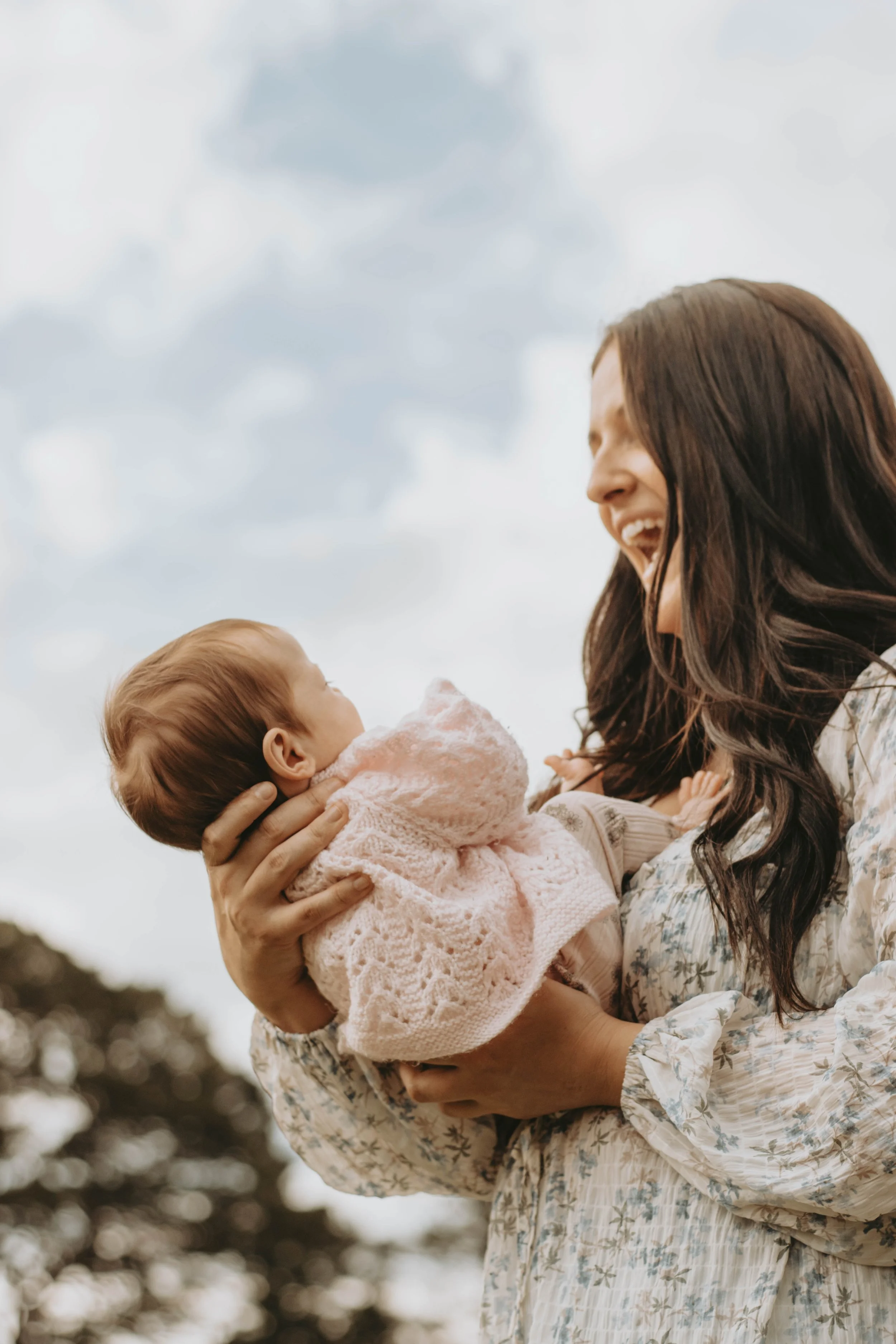 A woman holding a baby and smiling at each other outdoors against a cloudy sky.