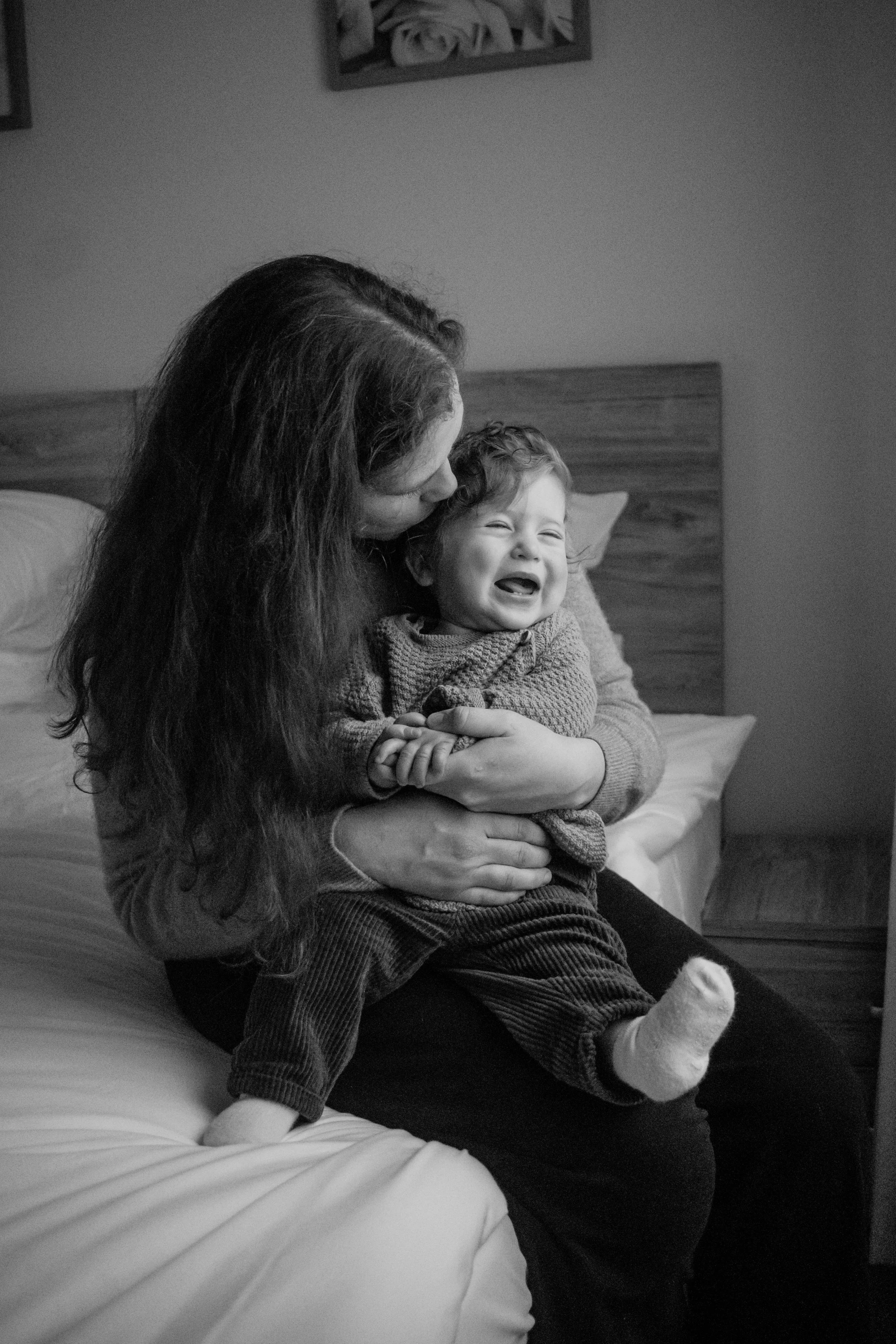 A woman with long dark hair is sitting on a bed, holding a young child with curly hair, both smiling and laughing. The woman is kissing the child's head, and the child is wearing a sweater and striped pants.