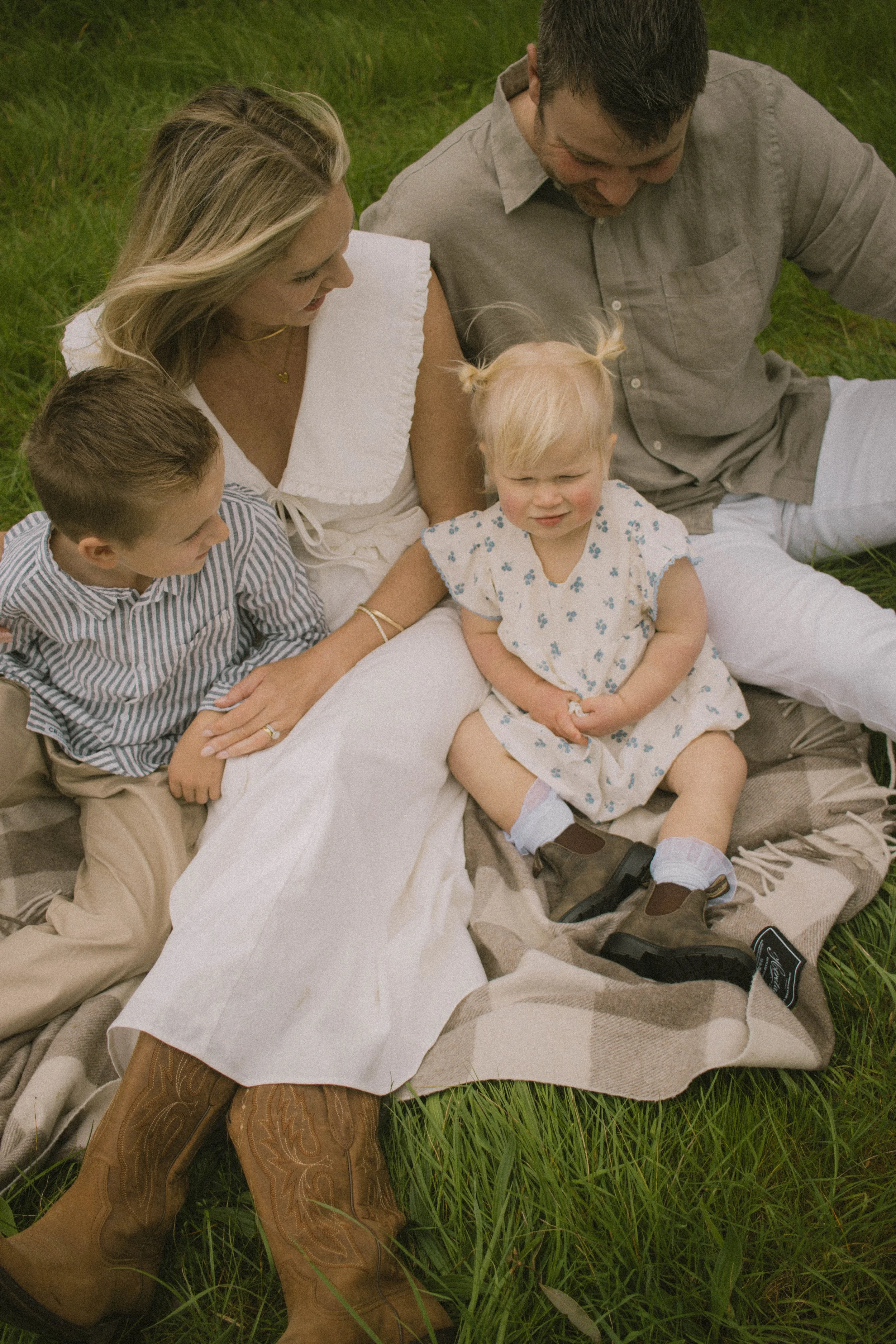 A family of five sitting and lying on a plaid blanket on green grass, with one woman, one man, three children, all dressed casually, enjoying outdoor time.