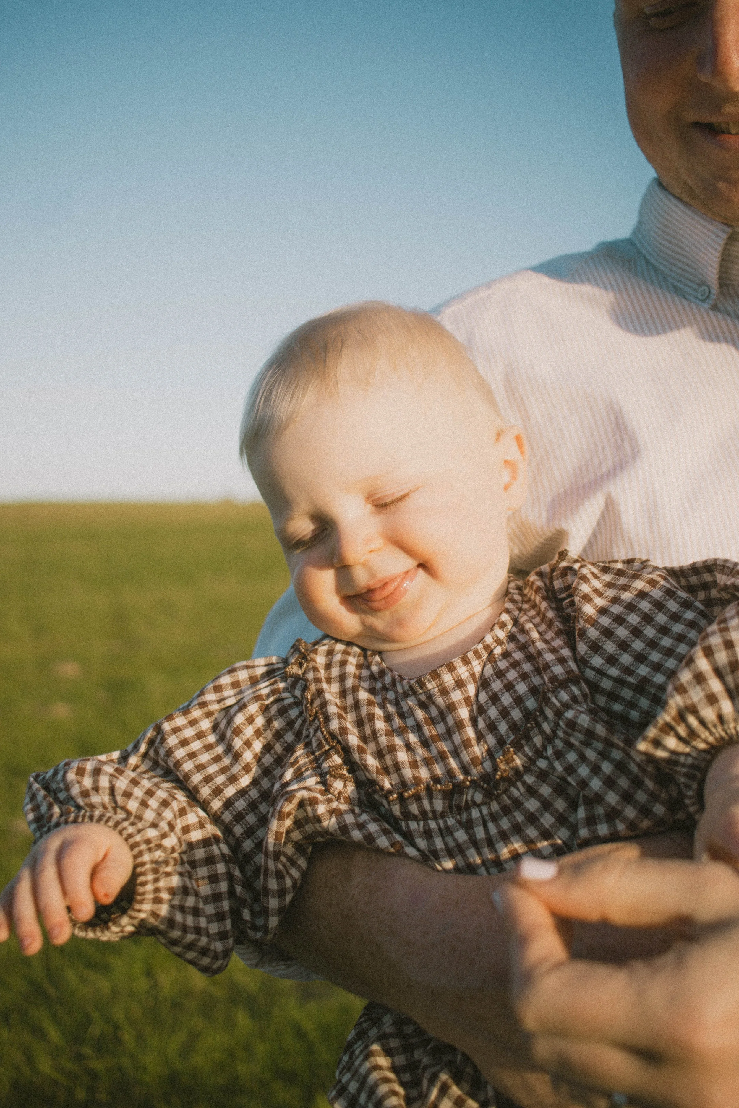 A young child with blonde hair smiling with eyes closed, wearing a brown checkered dress, being held by an adult woman outdoors in a field with a clear blue sky in the background.