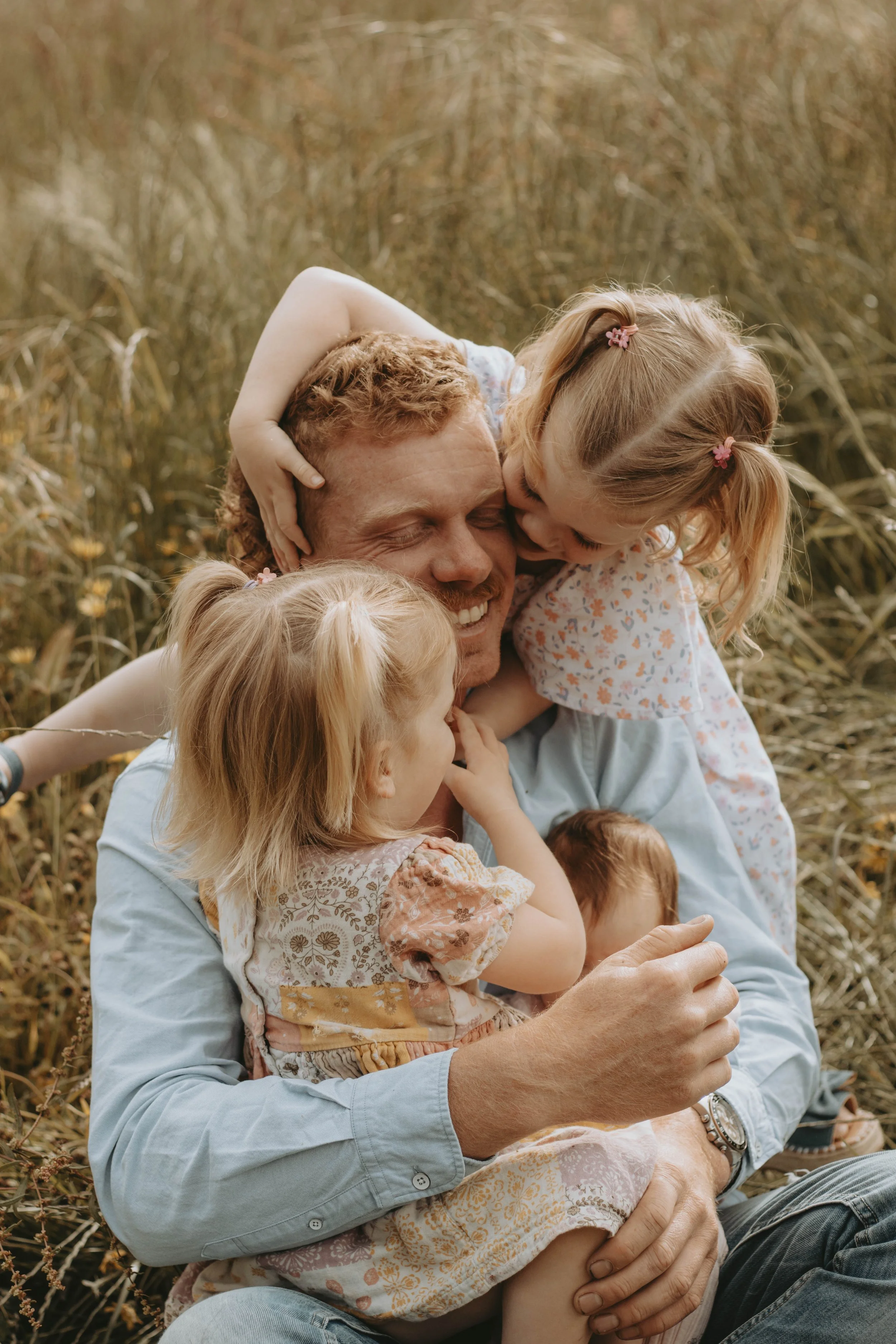 A man with three young girls hugging and playing together in a field of tall grass.