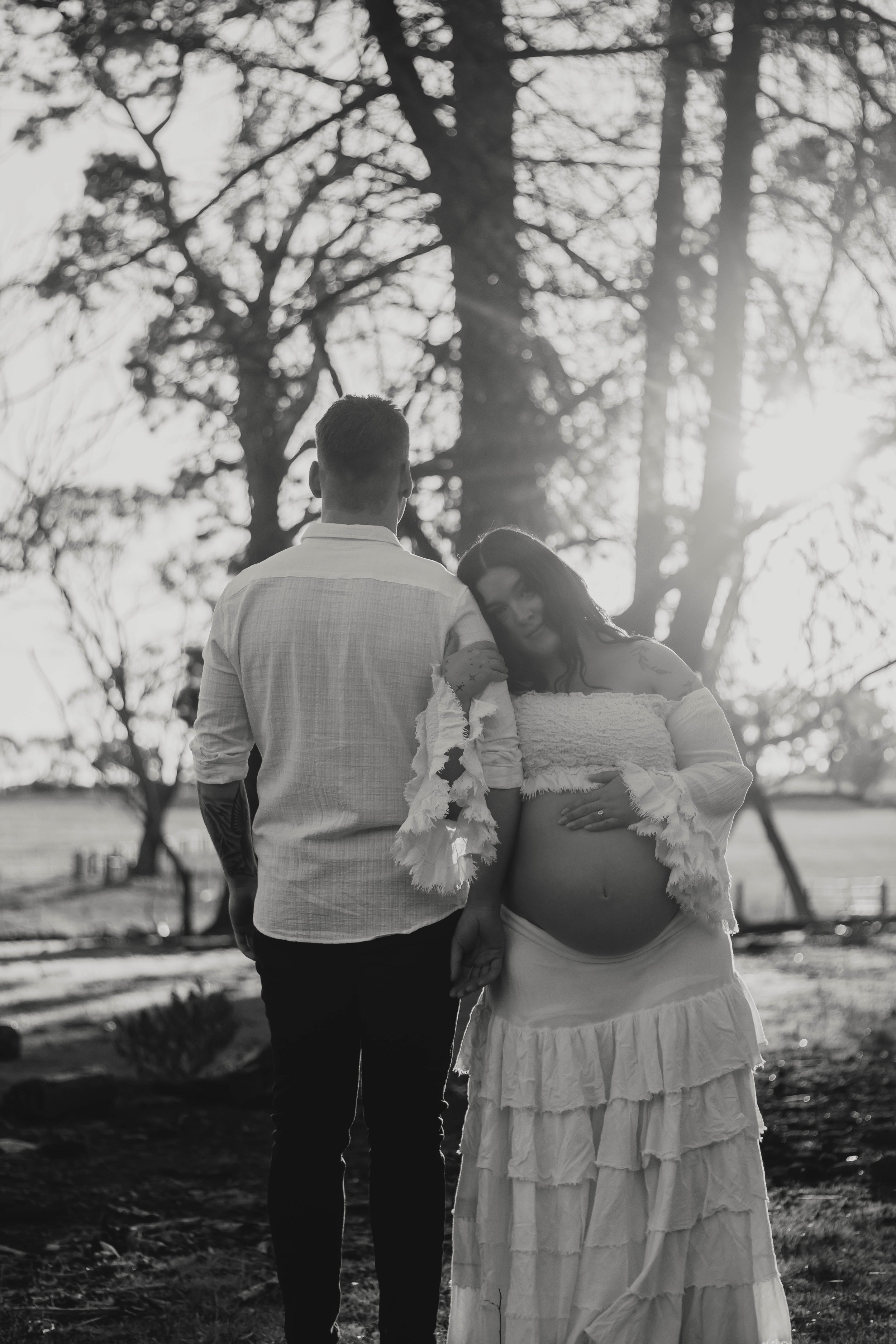 A couple standing outdoors in a park with trees in the background, the woman appears pregnant and is wearing a strapless dress, while the man stands beside her, both facing away from the camera, in black and white.