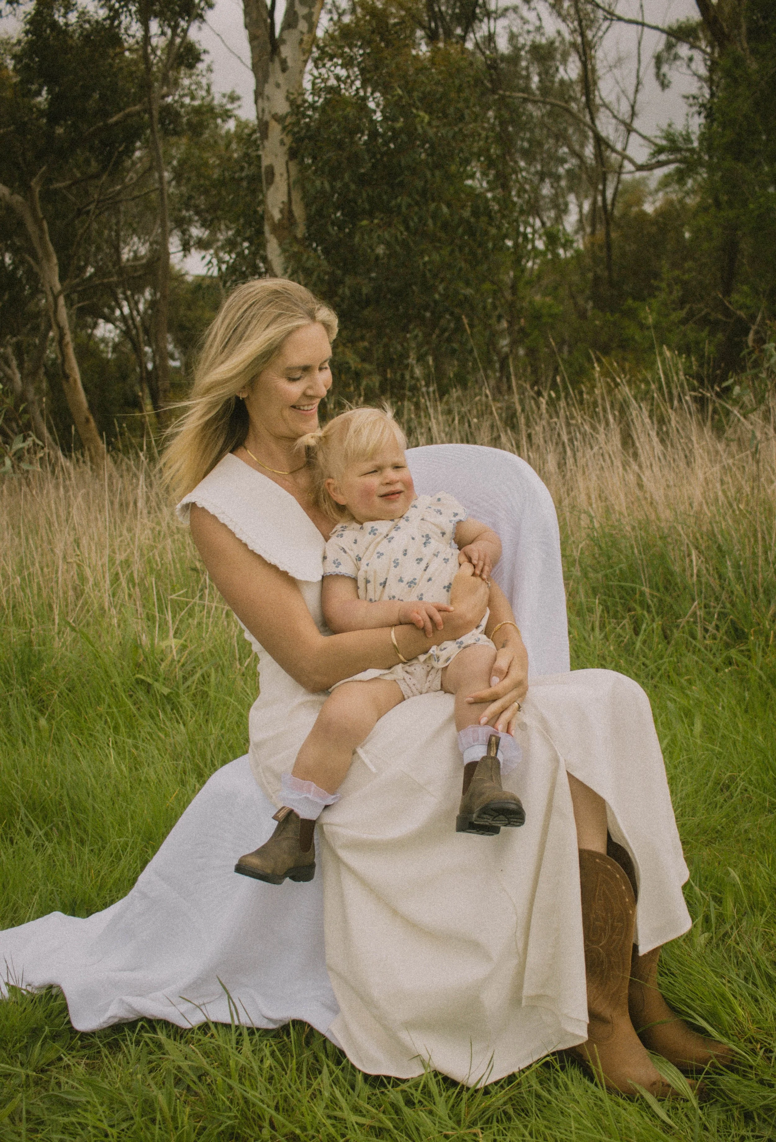 A woman with long blonde hair wearing a white dress sitting outdoors in a grassy field holding a laughing blonde girl in a floral dress on her lap. The girl is wearing brown boots and has her arms crossed. The background features trees and overcast sky.