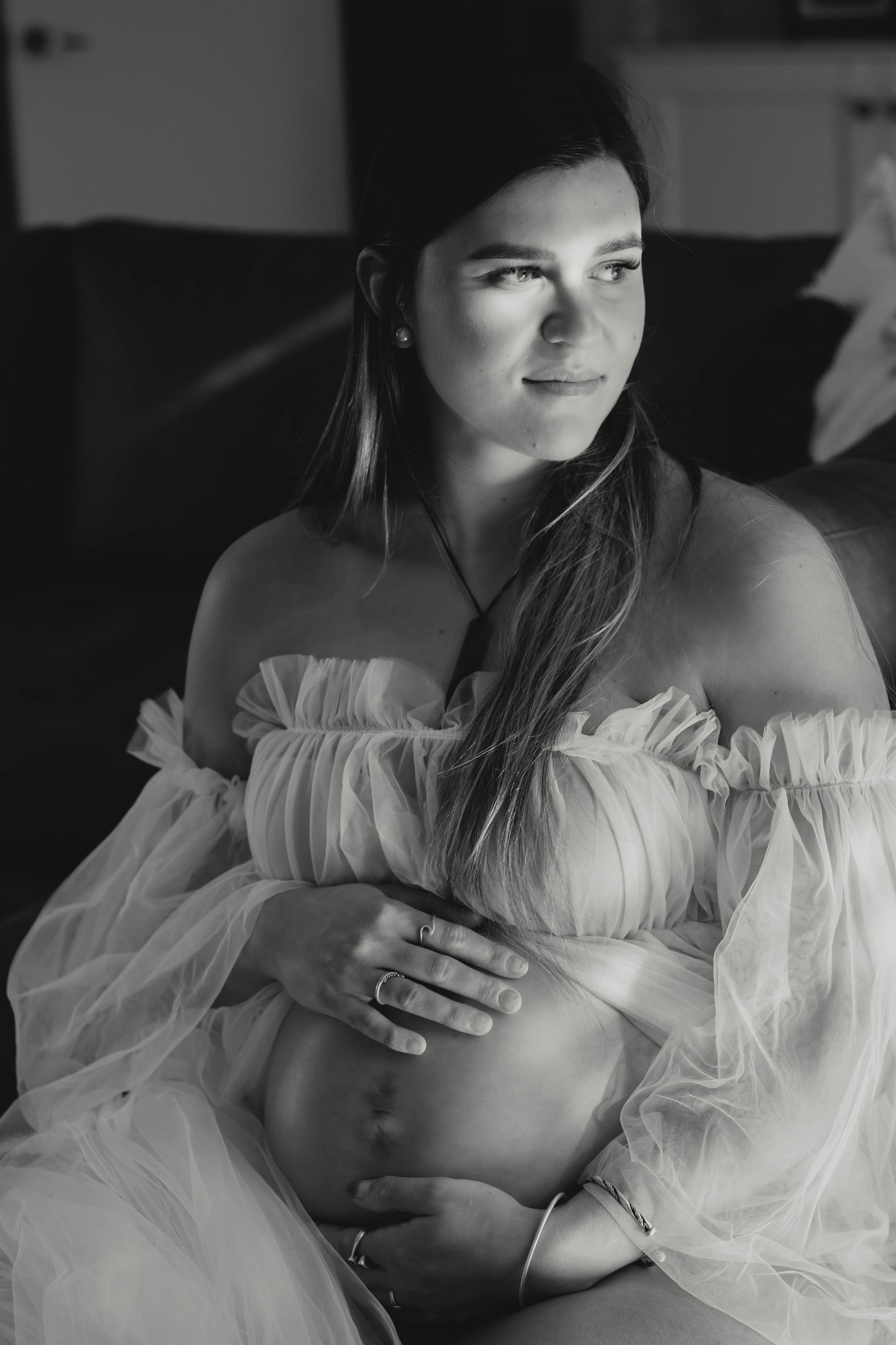 A woman with long hair, wearing an off-shoulder dress with ruffled sleeves, is sitting indoors, looking to her right with a thoughtful expression. She has her arms crossed over her chest, with her right hand resting on her left arm and her left hand 