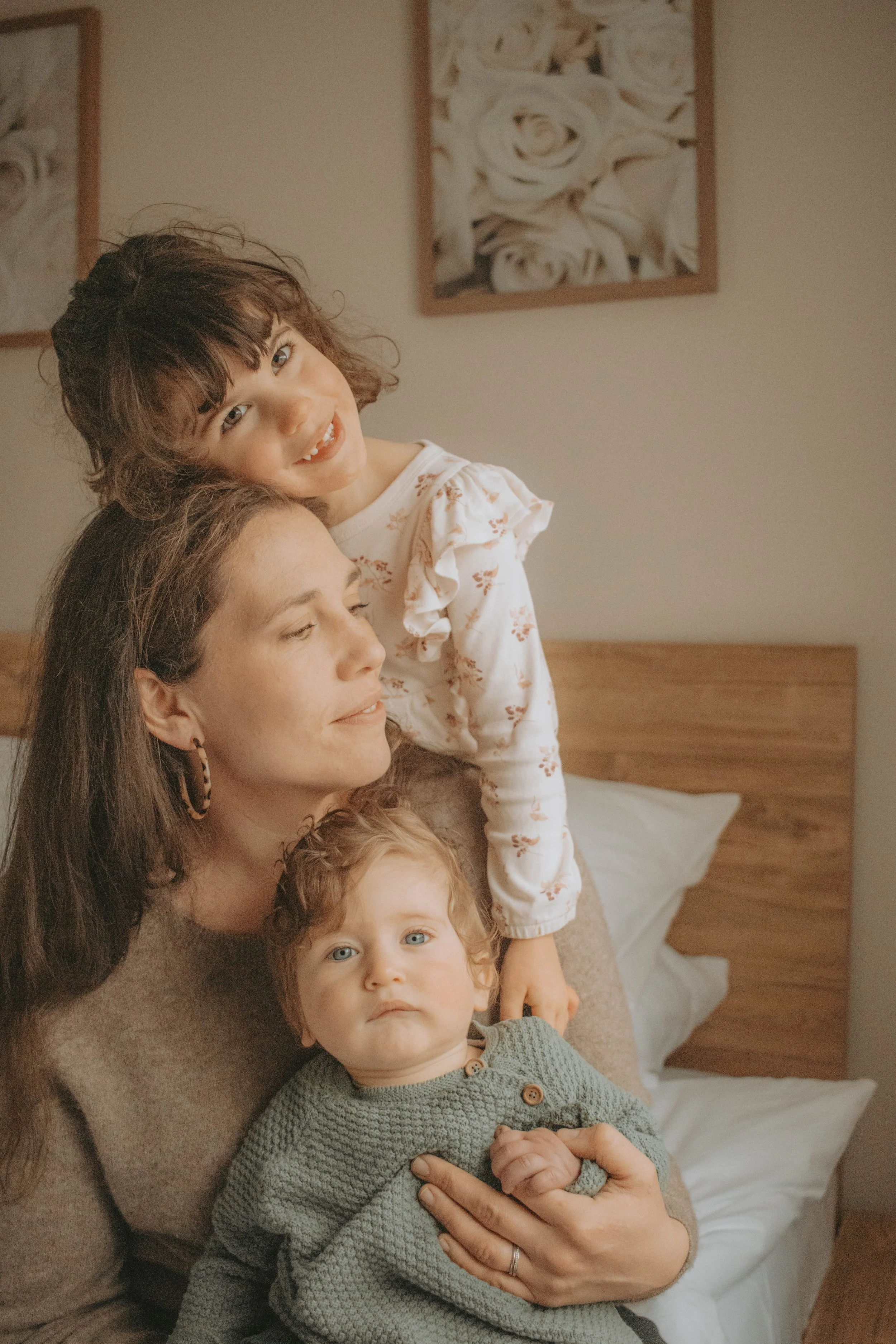 A woman with long brown hair holds a young boy with curly red hair, and a girl with brown hair and blue eyes is sitting on her lap. They are on a bed with a wooden headboard in a room with beige walls and framed artwork of white roses in the background.