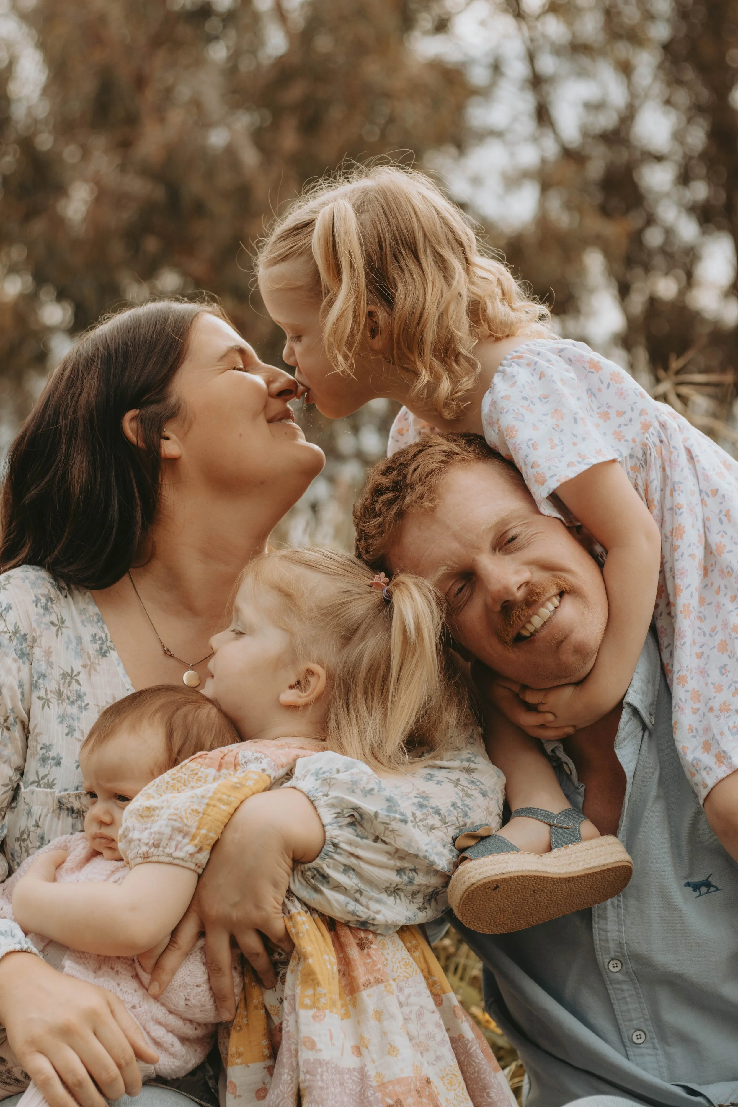 A family of five, including two women, one man, and three young children, enjoying a moment outdoors. The children are on the adults' shoulders, and everyone is smiling and showing affection.