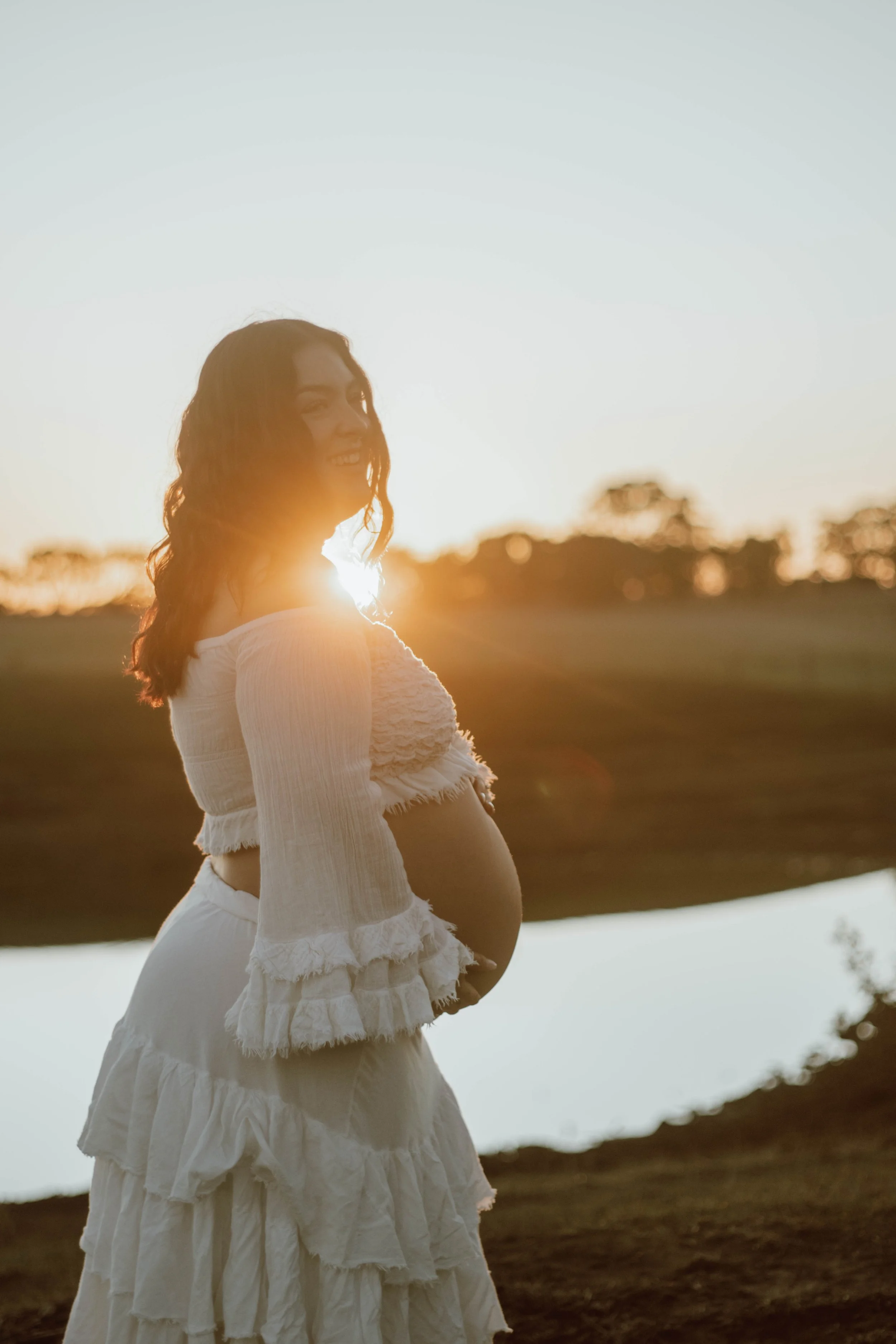 A pregnant woman wearing white clothing standing outdoors near a body of water, smiling, with the sun setting behind her and trees in the distance.