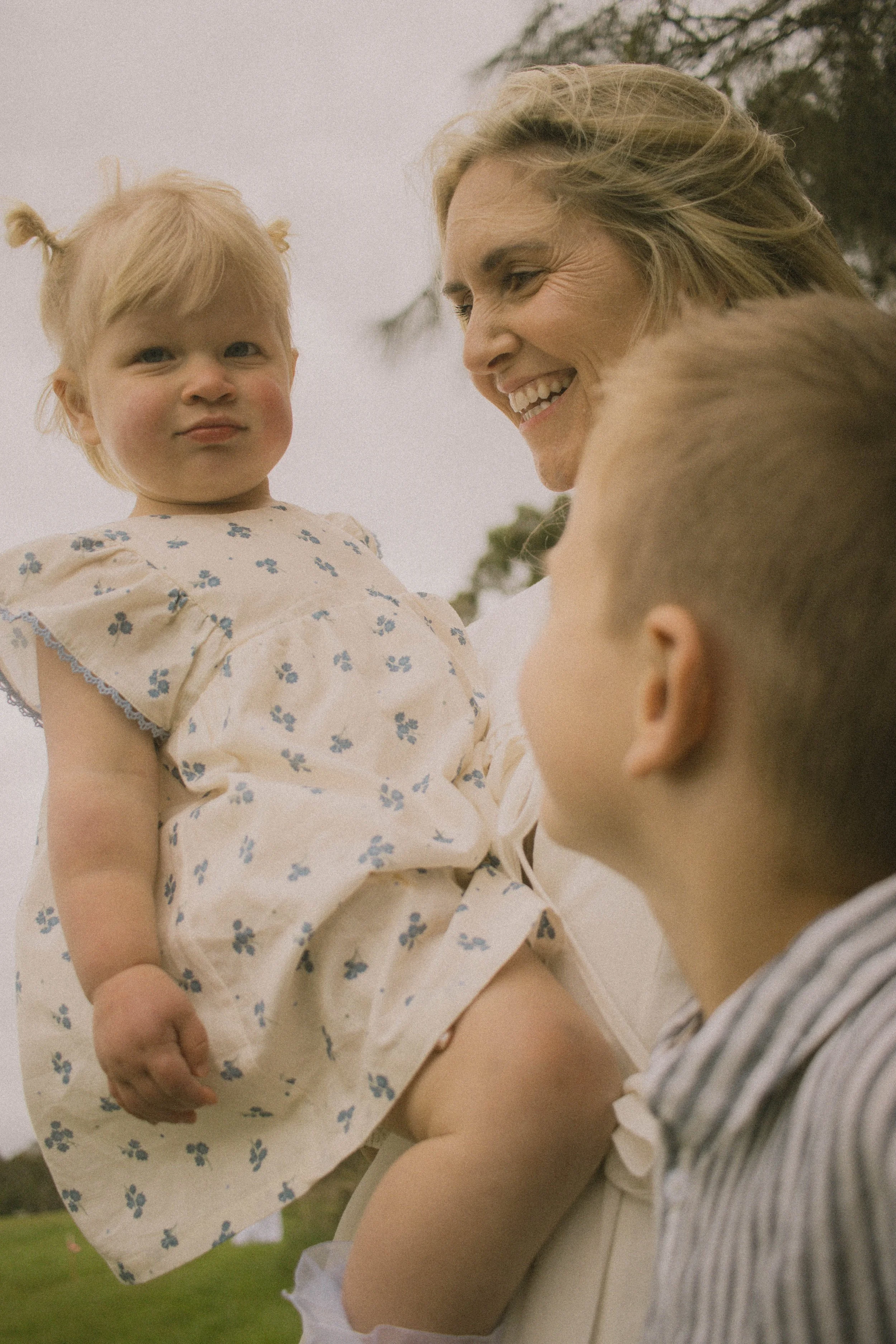 A woman holding a young girl outdoors with trees and cloudy sky in the background.
