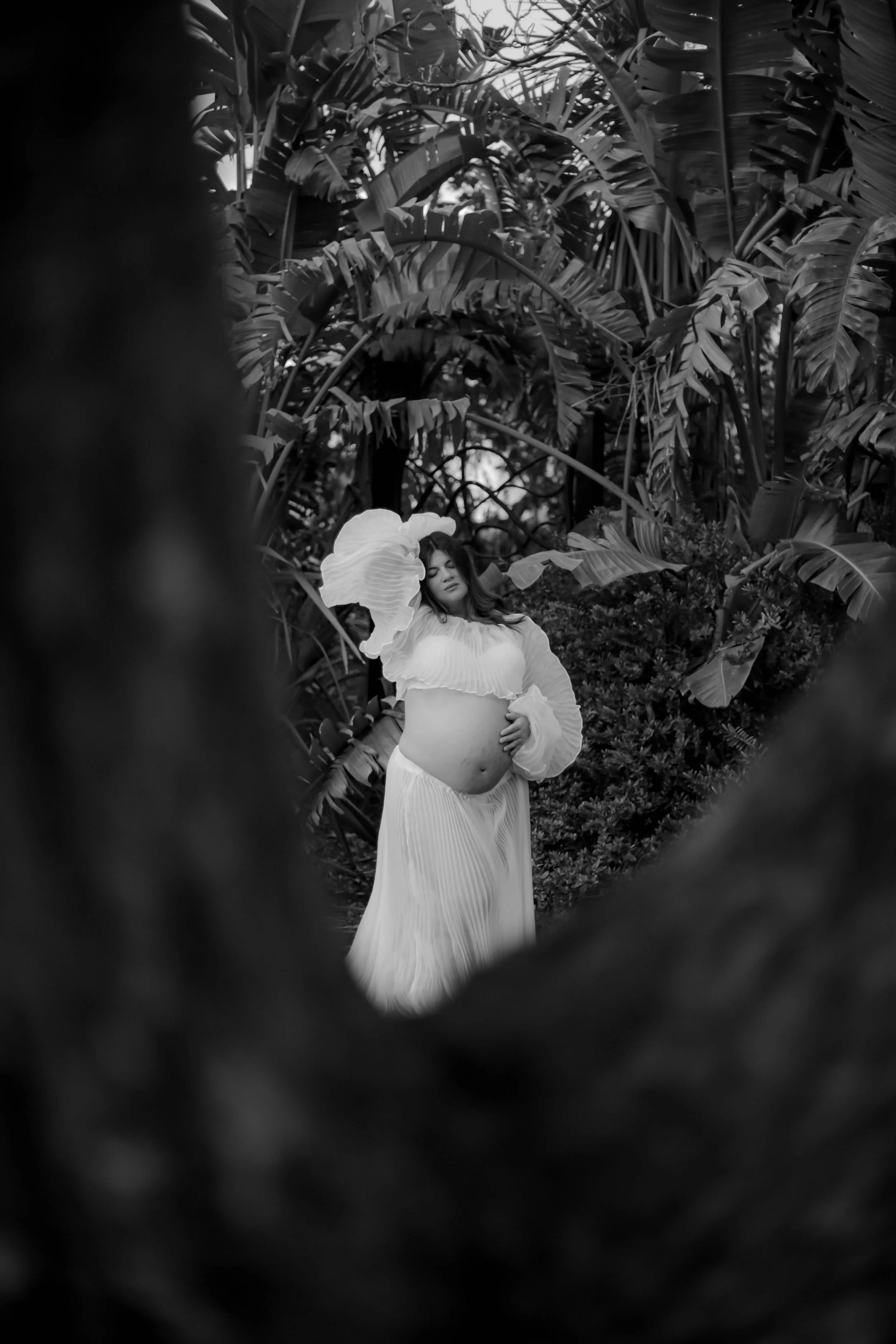 A woman in a flowing white dress and a wide-brimmed hat posing among dense tropical plants in a black and white photograph.