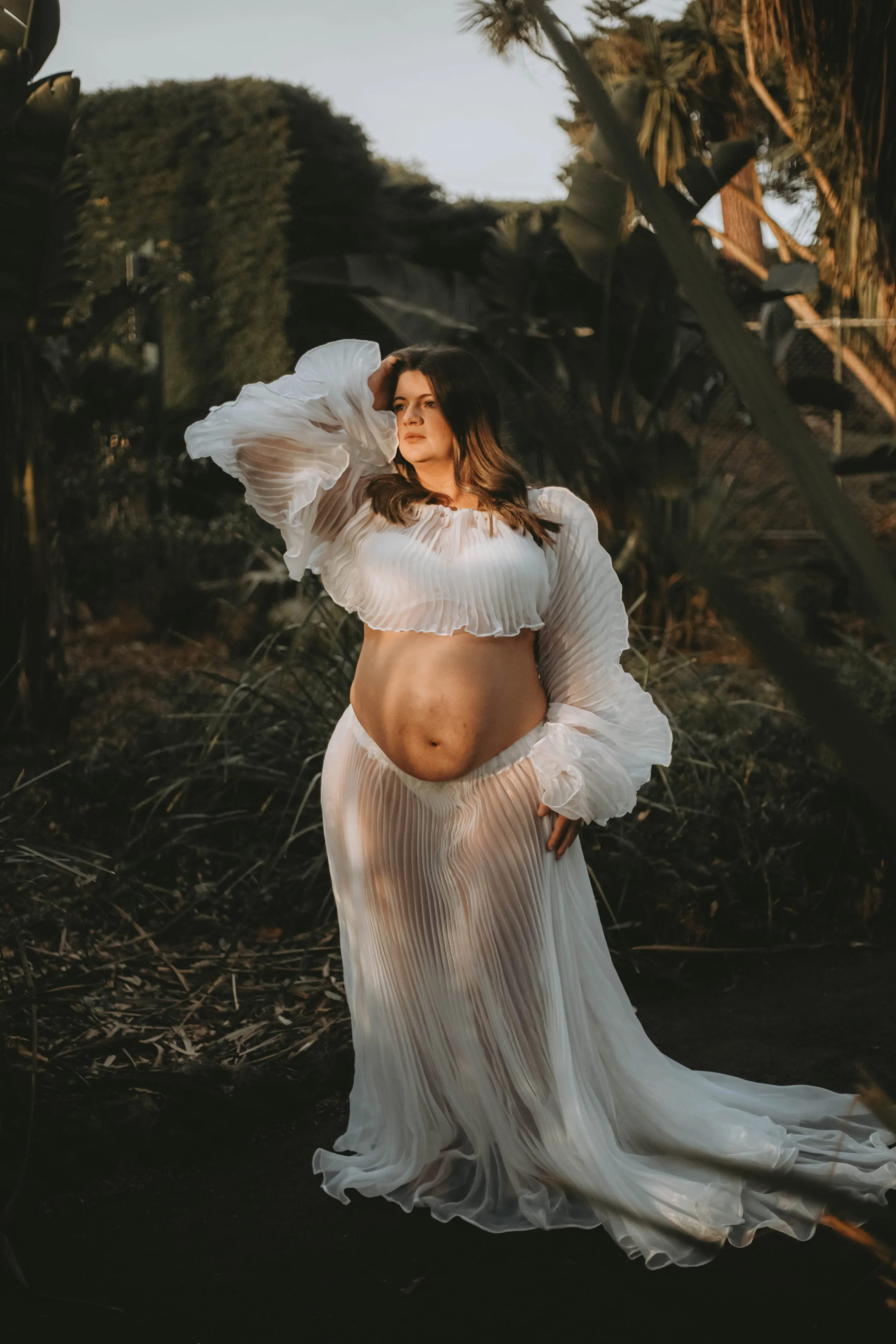 A woman wearing a flowing, pleated white dress with long, ruffled sleeves standing outdoors among trees and plants at dusk.