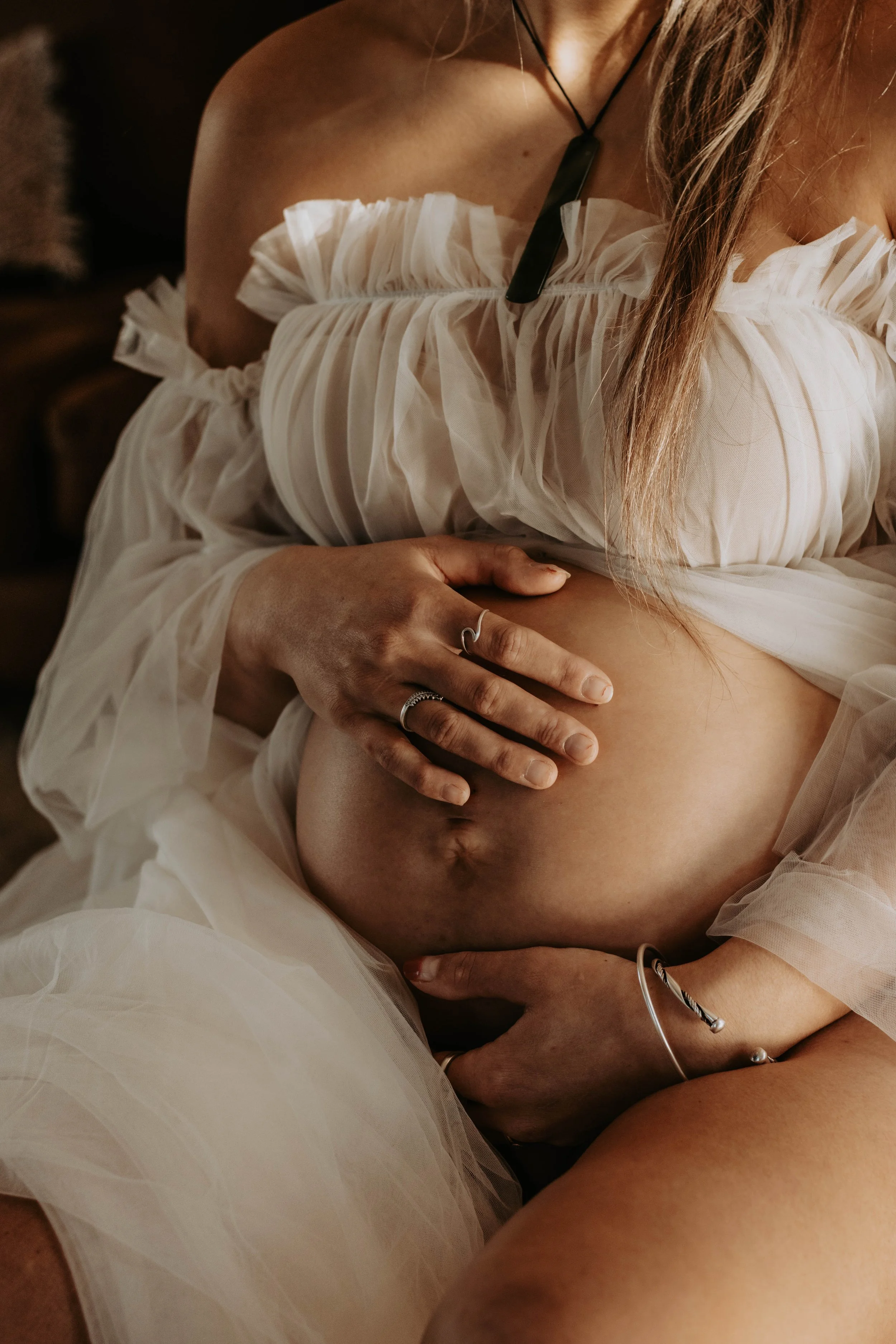 A close-up of a woman's torso wearing a white, ruffled off-shoulder dress. The woman's hand is resting on her bare stomach, with jewelry including rings, a bracelet, and a necklace visible. The setting appears intimate with warm lighting.