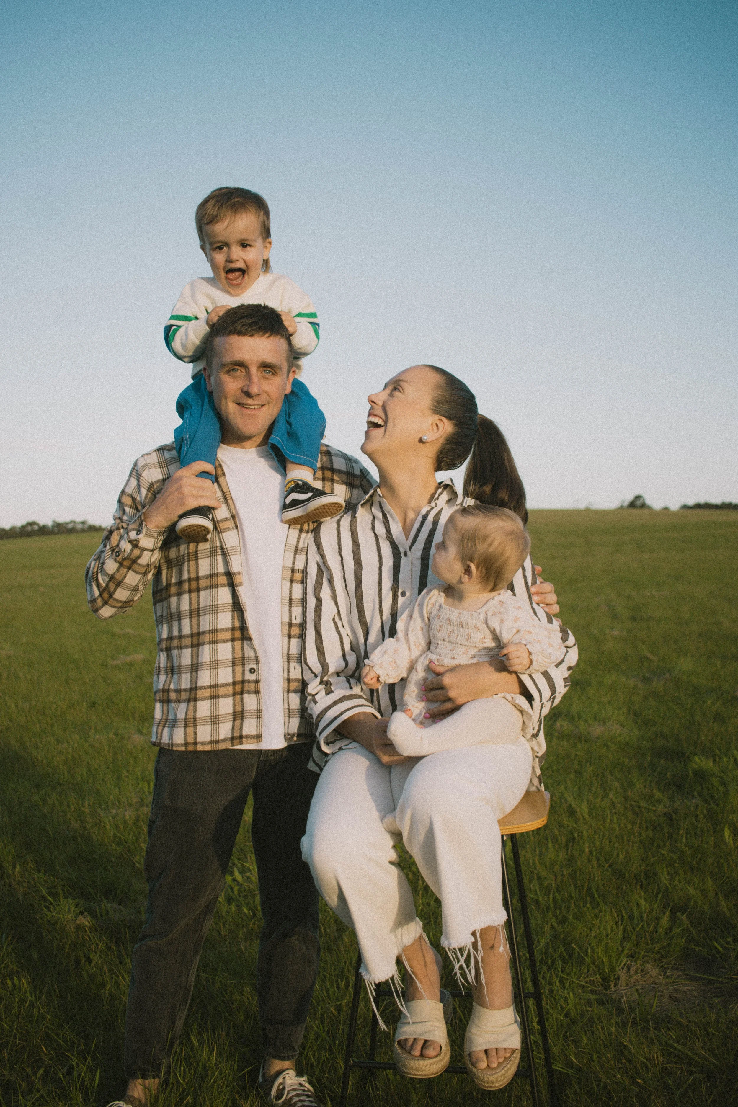 A family of four enjoying time outdoors in a grassy field during the daytime. The father is standing, carrying a young boy on his shoulders, who is smiling with his mouth open. The mother is sitting on a stool, holding a young girl on her lap, both l
