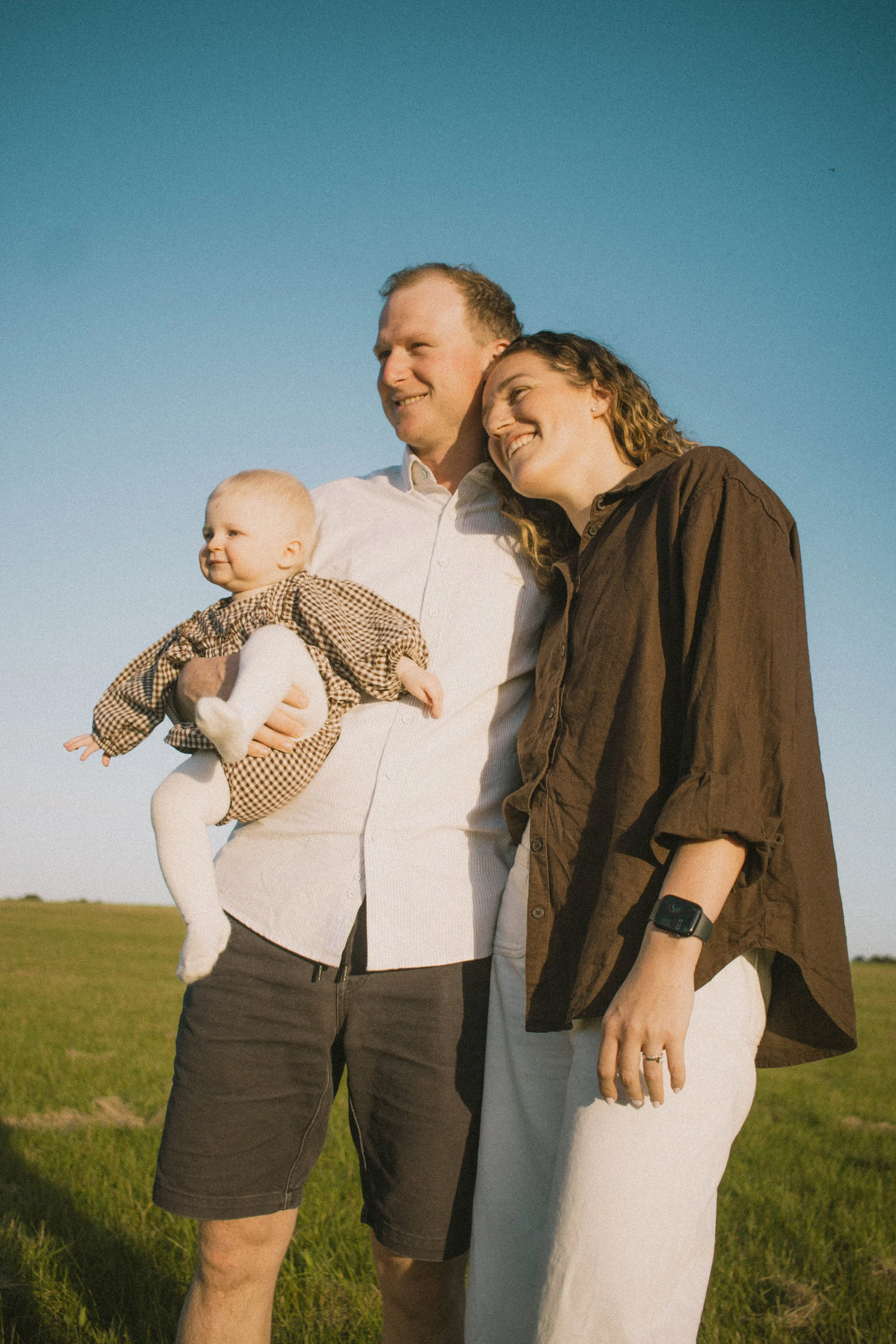 A family of three standing outdoors on a grassy field with clear blue sky, smiling and enjoying the moment. The man is holding a baby girl, and a woman stands close with her head leaning on the man's shoulder.