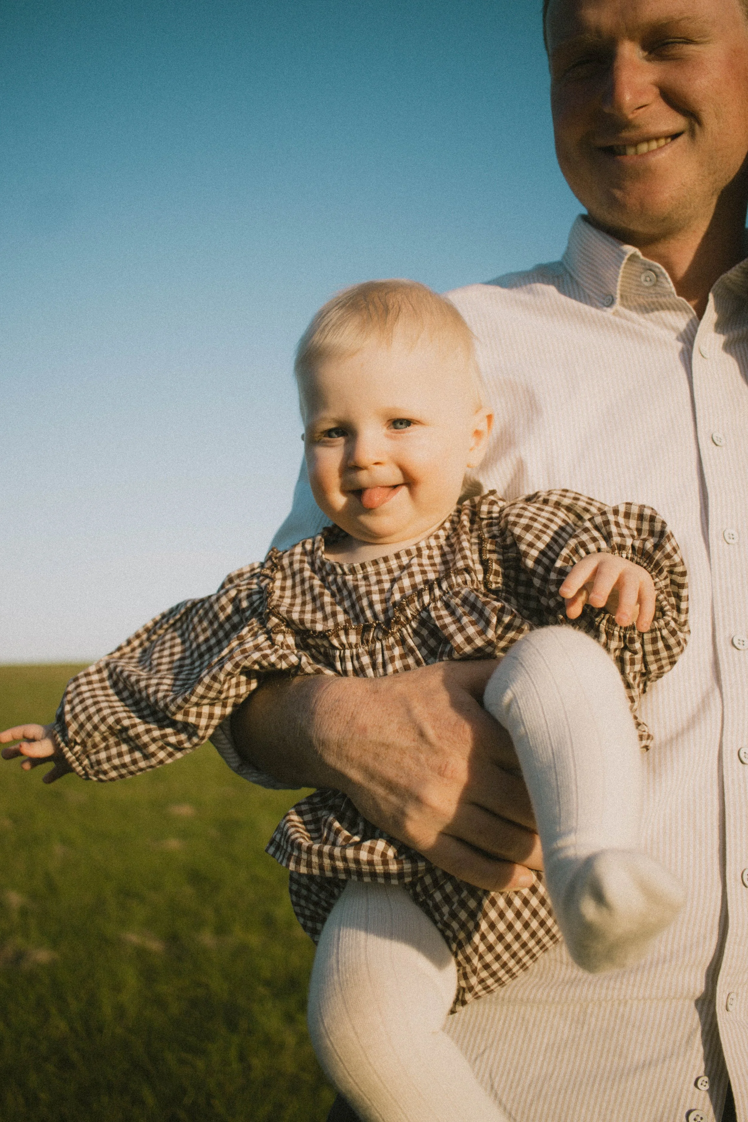 A man is holding a happy young girl outdoors in a grassy area during daylight. The girl is sticking out her tongue and wearing a checkered dress, while the man is smiling.