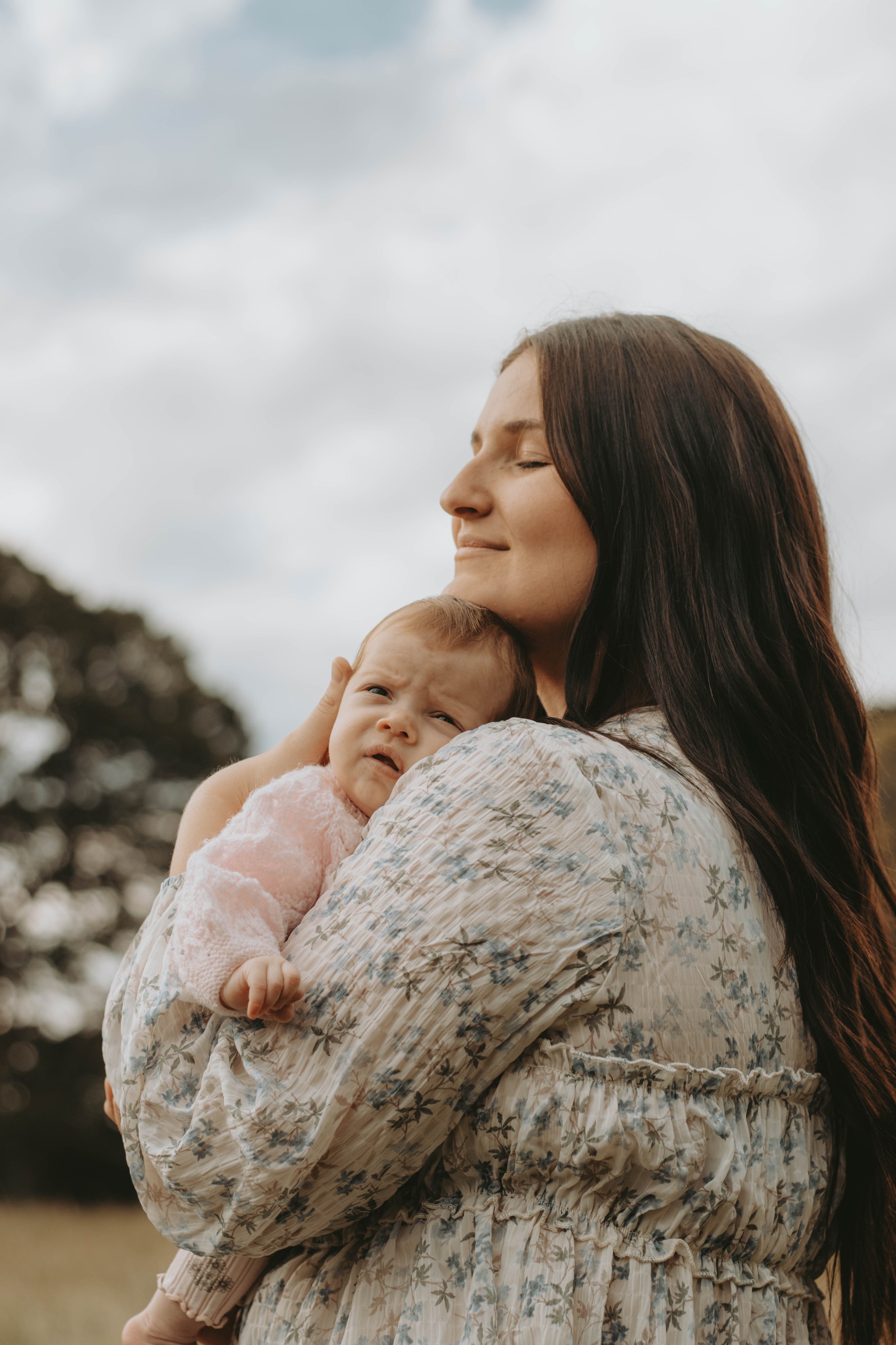 A woman with long brown hair holding a baby in a pink outfit against a cloudy sky and trees in the background.