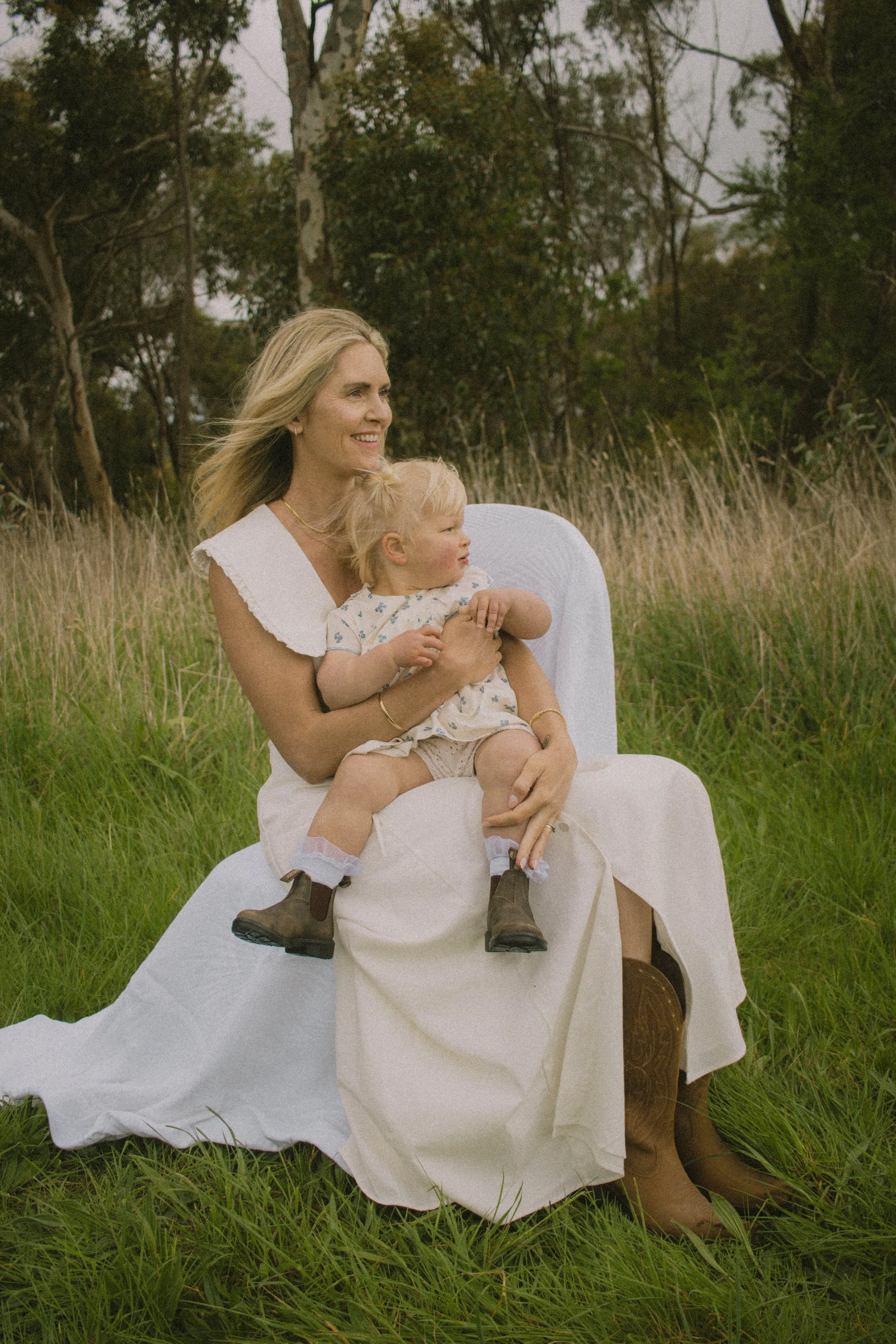 A woman with long blonde hair sitting in a field holding a young girl with blonde hair. Both are smiling, surrounded by trees and tall grass, wearing casual summer clothes.