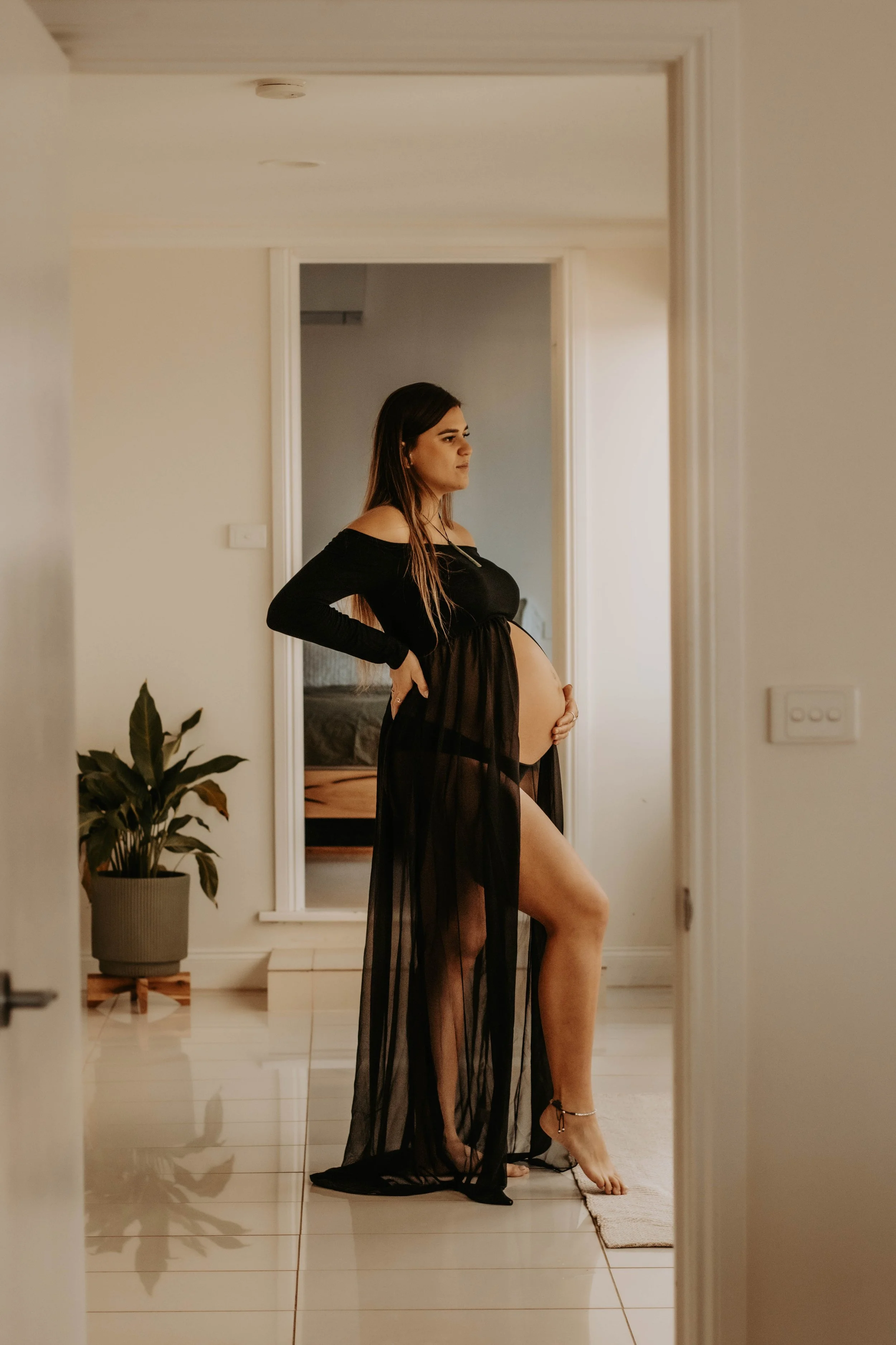 A pregnant woman is standing in a doorway, wearing black sheer lingerie with off-shoulder sleeves and high heels, with her left hand on her hip and her right hand resting on her belly, inside a home with white walls, white tiled floor, a potted plant, and a mirror reflecting a bedroom in the background.