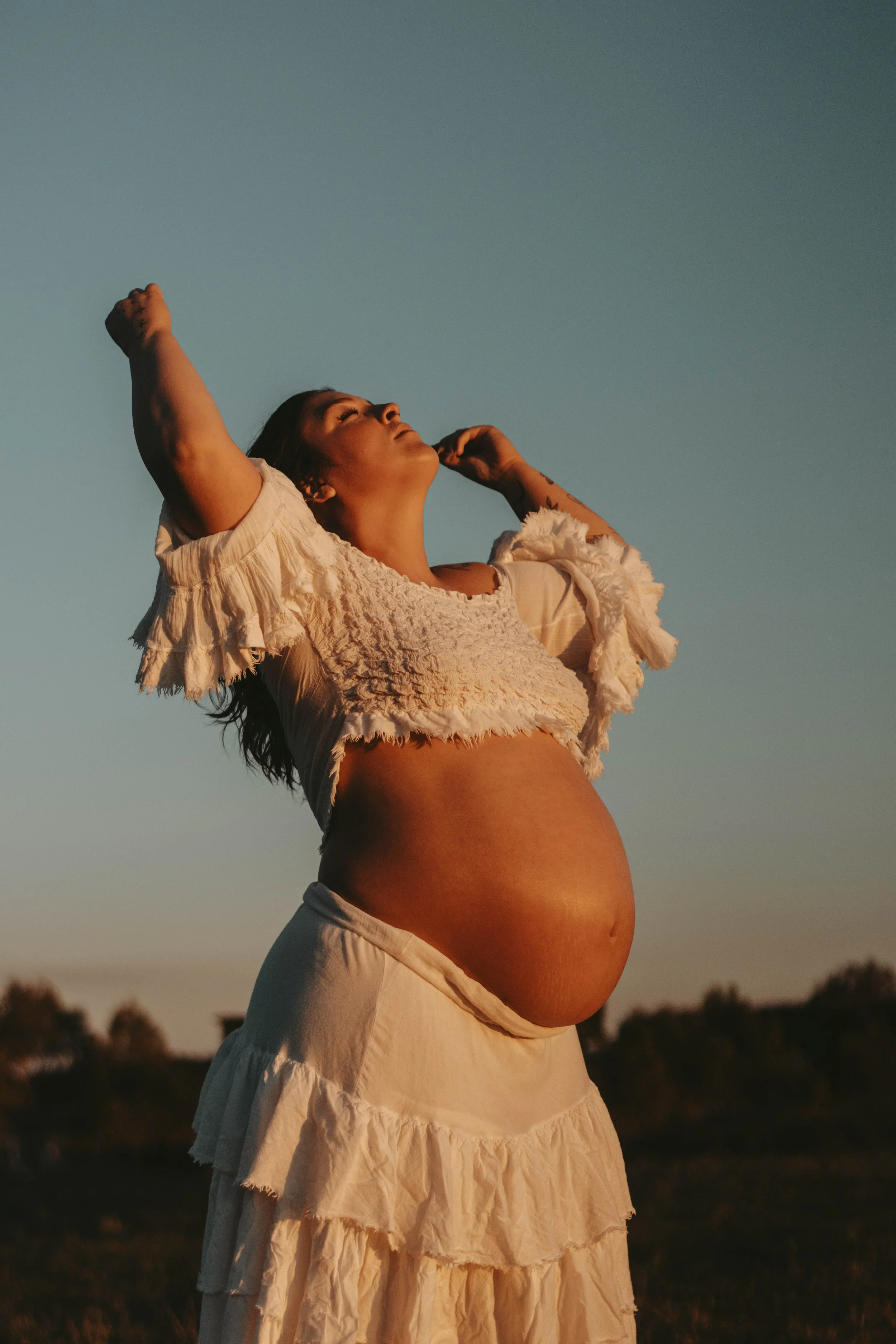 A pregnant woman with dark hair wearing a white ruffled top and skirt, standing outdoors at sunset with her arms raised and eyes closed.