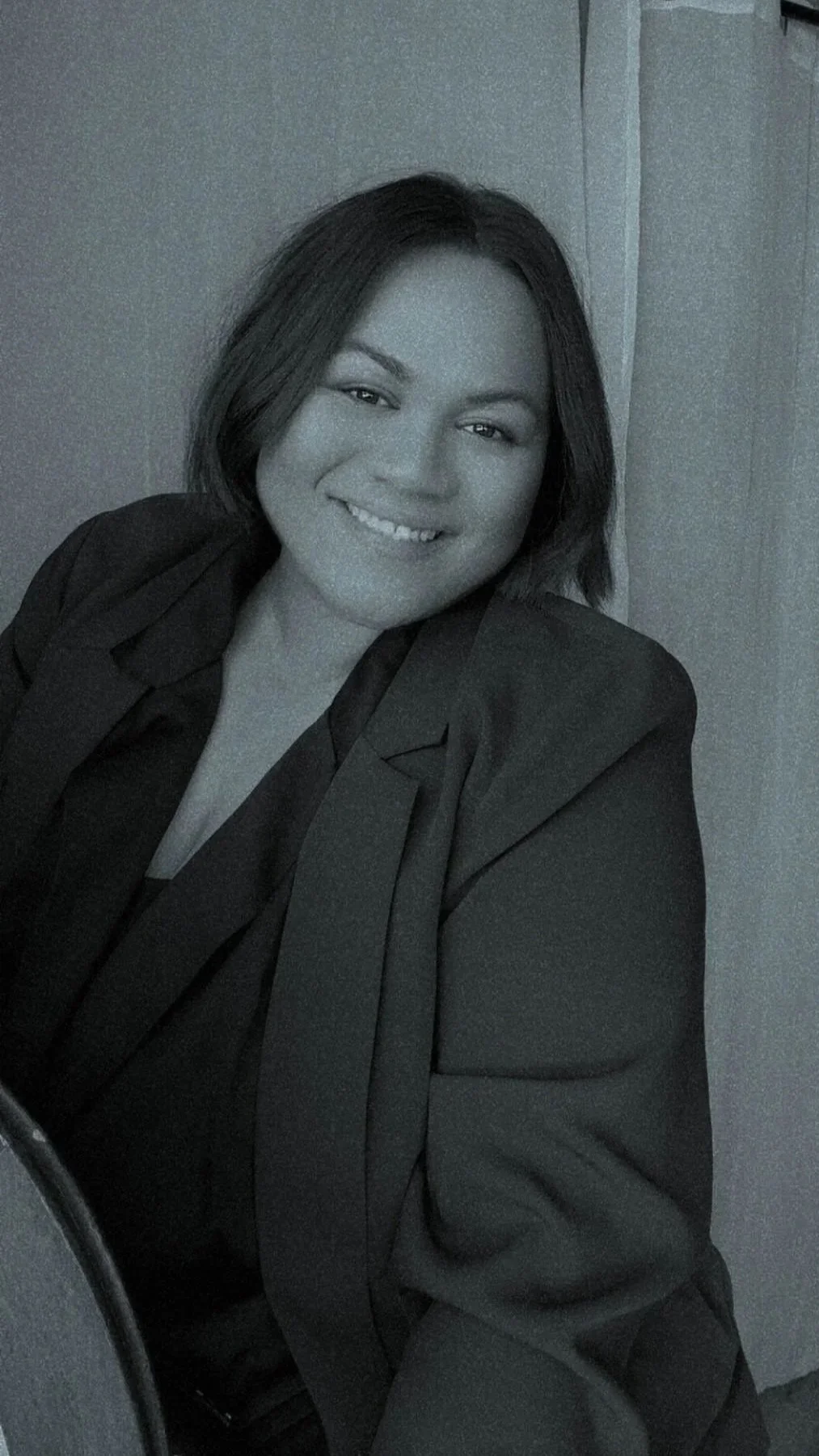 Black and white photo of a woman with shoulder-length dark hair, smiling, wearing a dark blazer and gloves, sitting in front of a plain wall.