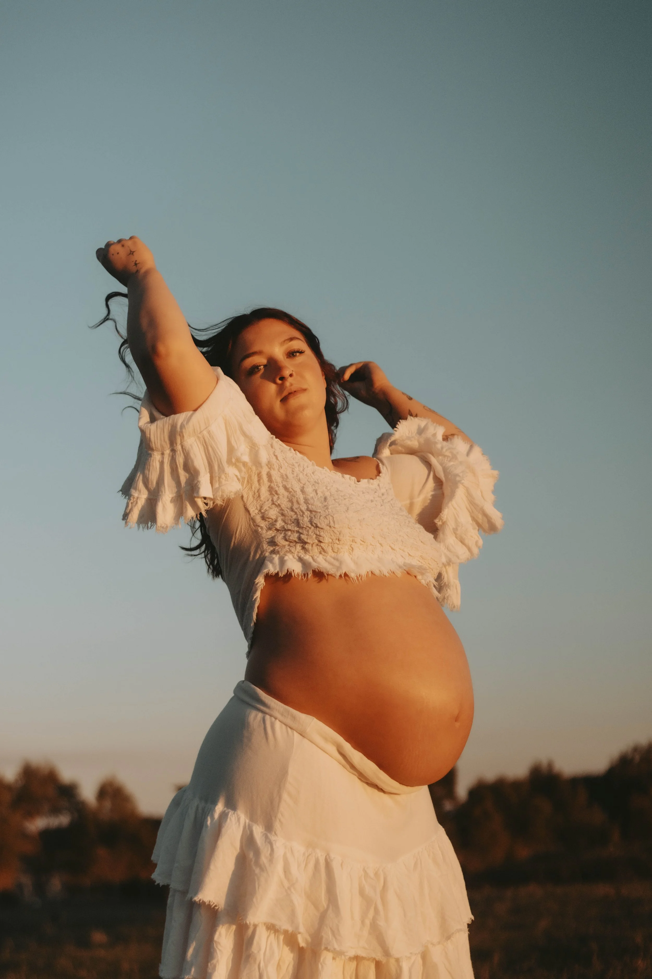 A pregnant woman in a white ruffled top and skirt standing outdoors during sunset, with one arm raised above her head and the other touching her hair, against a backdrop of trees and a clear sky.