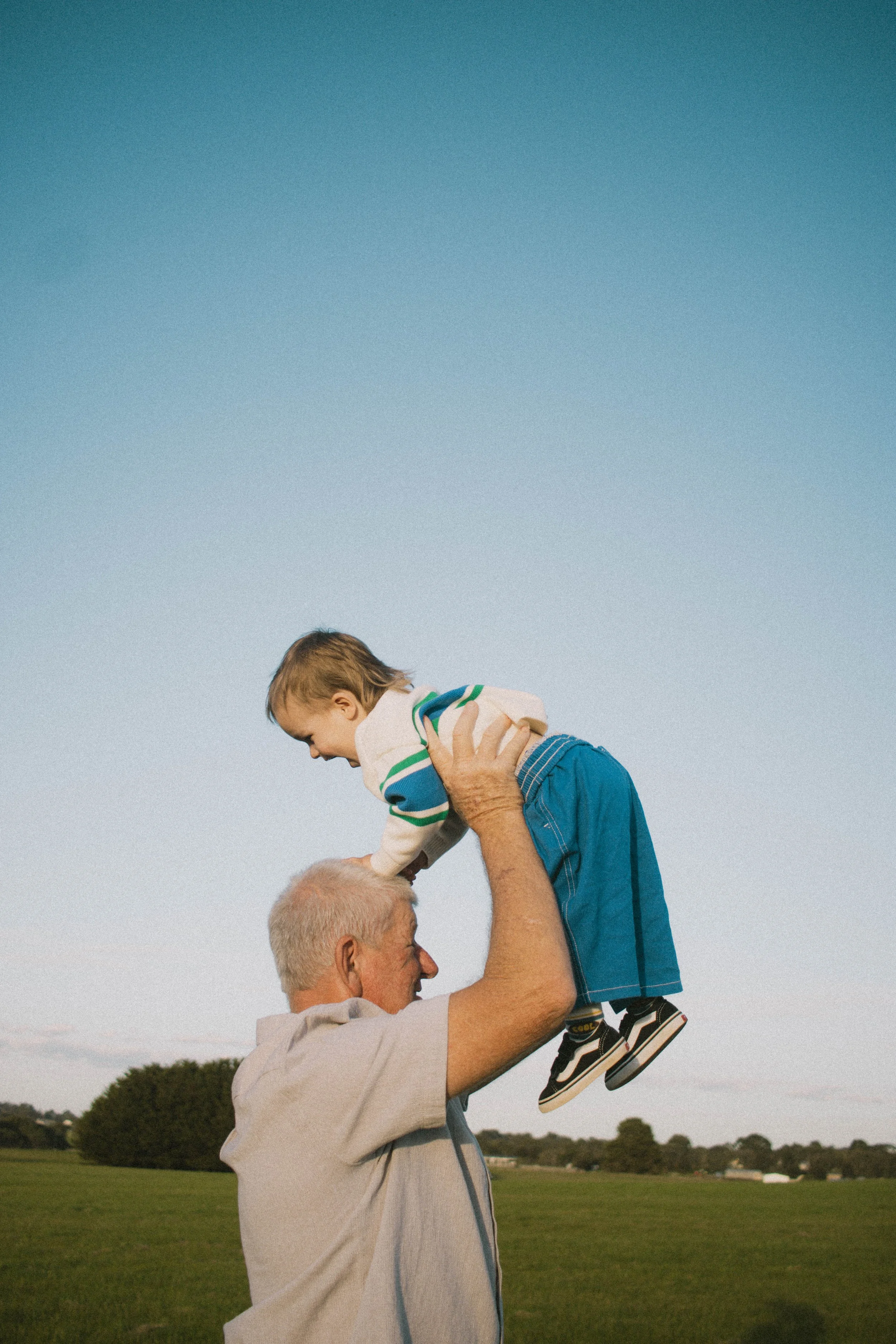 An elderly man lifting a young boy into the air in an outdoor park with a clear blue sky and trees in the background.