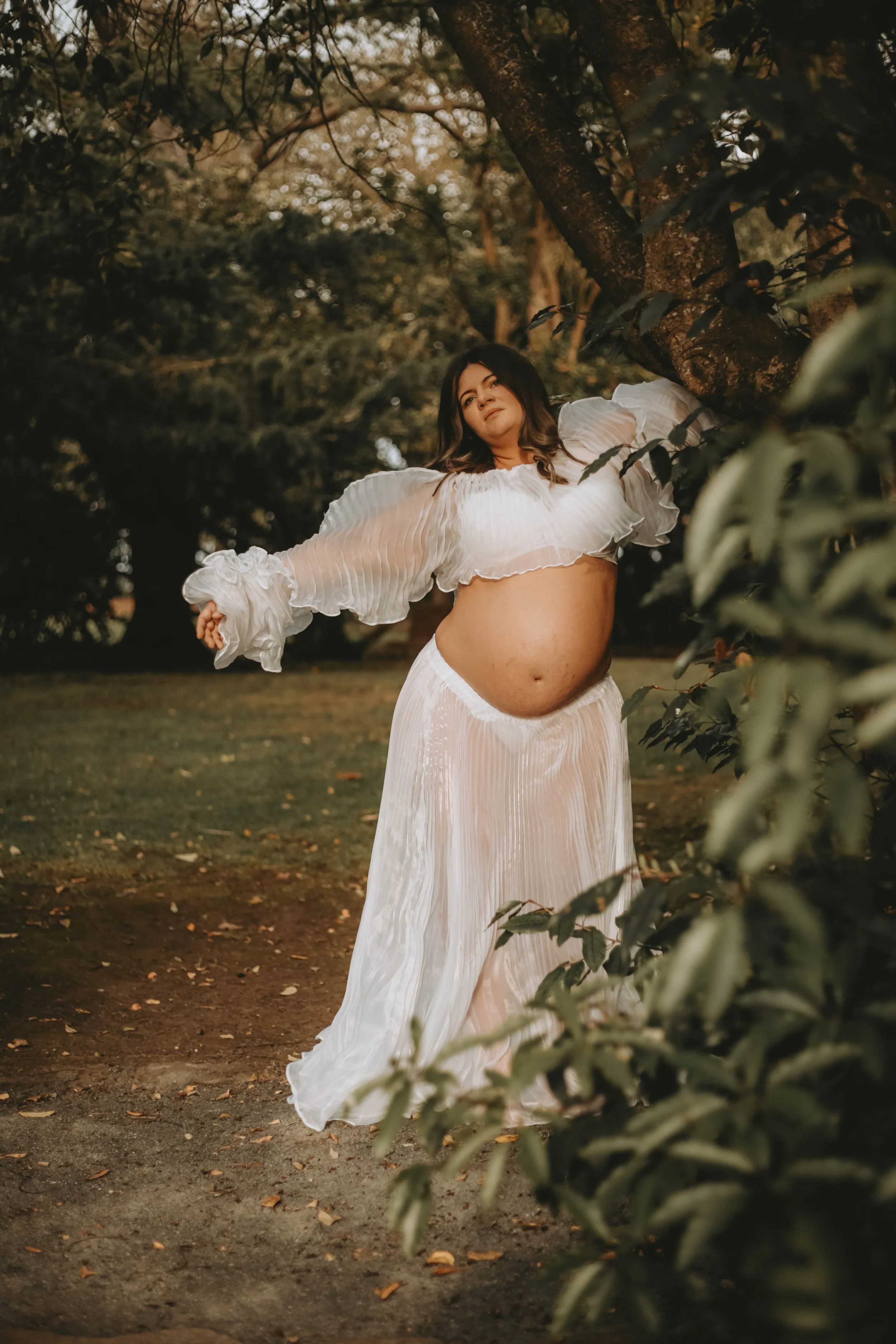 A pregnant woman in a flowing white dress standing outdoors near a tree, looking thoughtfully into the distance amid greenery and dappled sunlight.