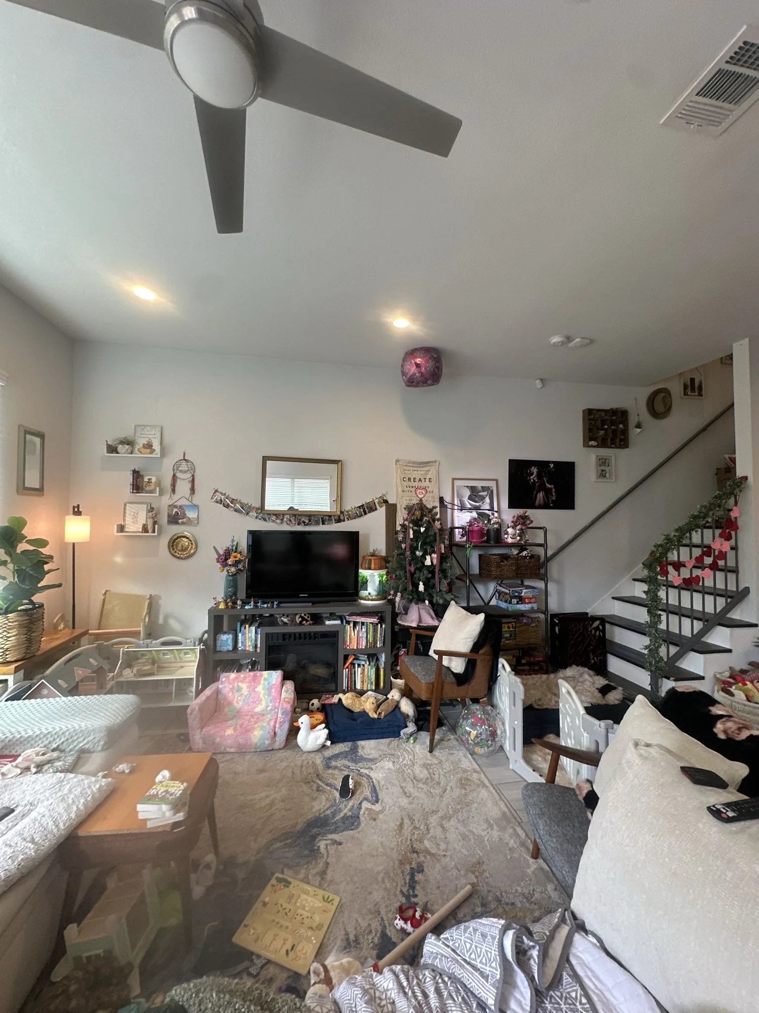 A cluttered living room with furniture and various items scattered around, including a television, chairs, a coffee table, and stuffed animals on the floor.