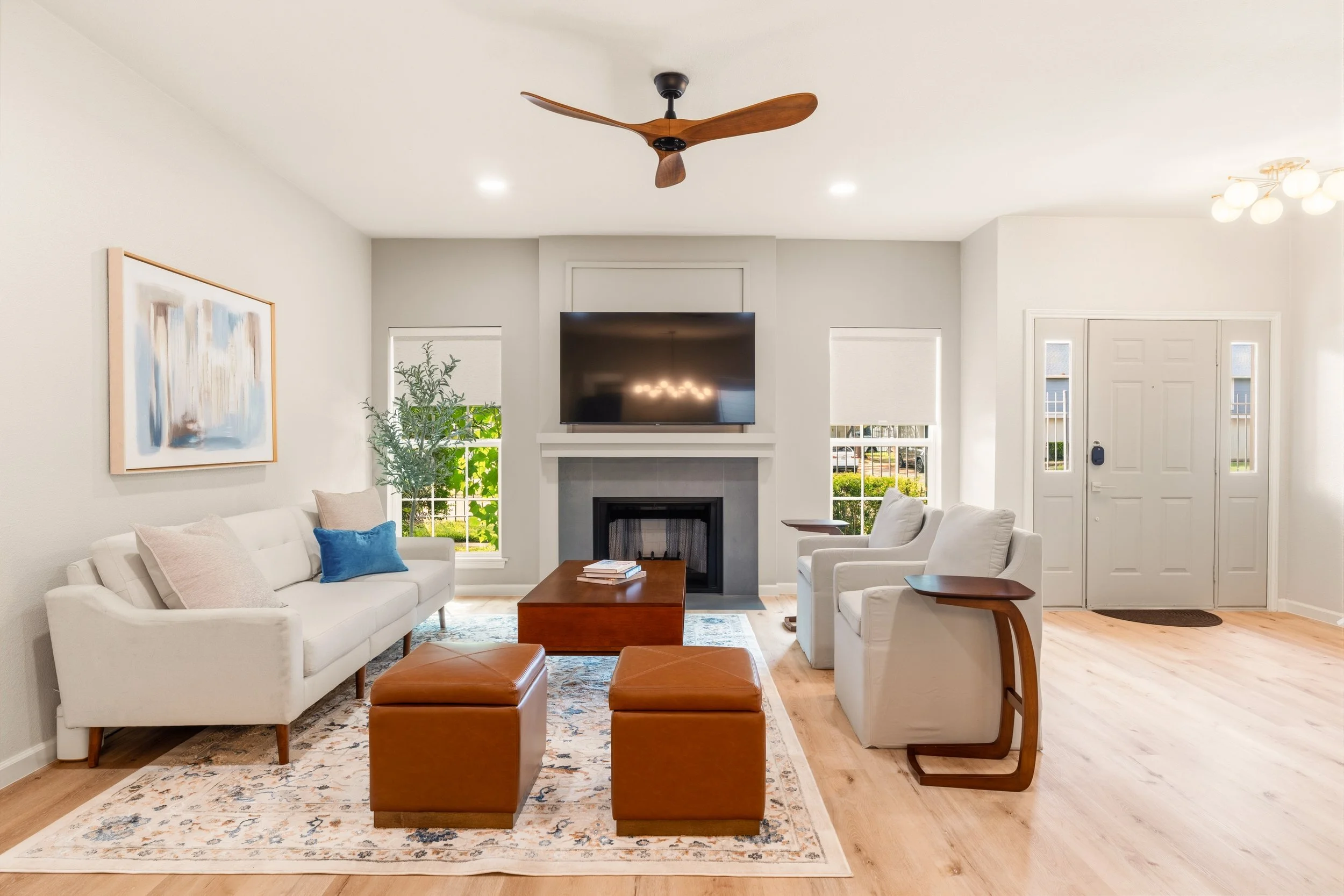 Living room with white sofa, two orange ottomans, white armchair, coffee table, fireplace, mounted TV, abstract wall art, potted plant, hardwood floor, ceiling fan, windows, front door.