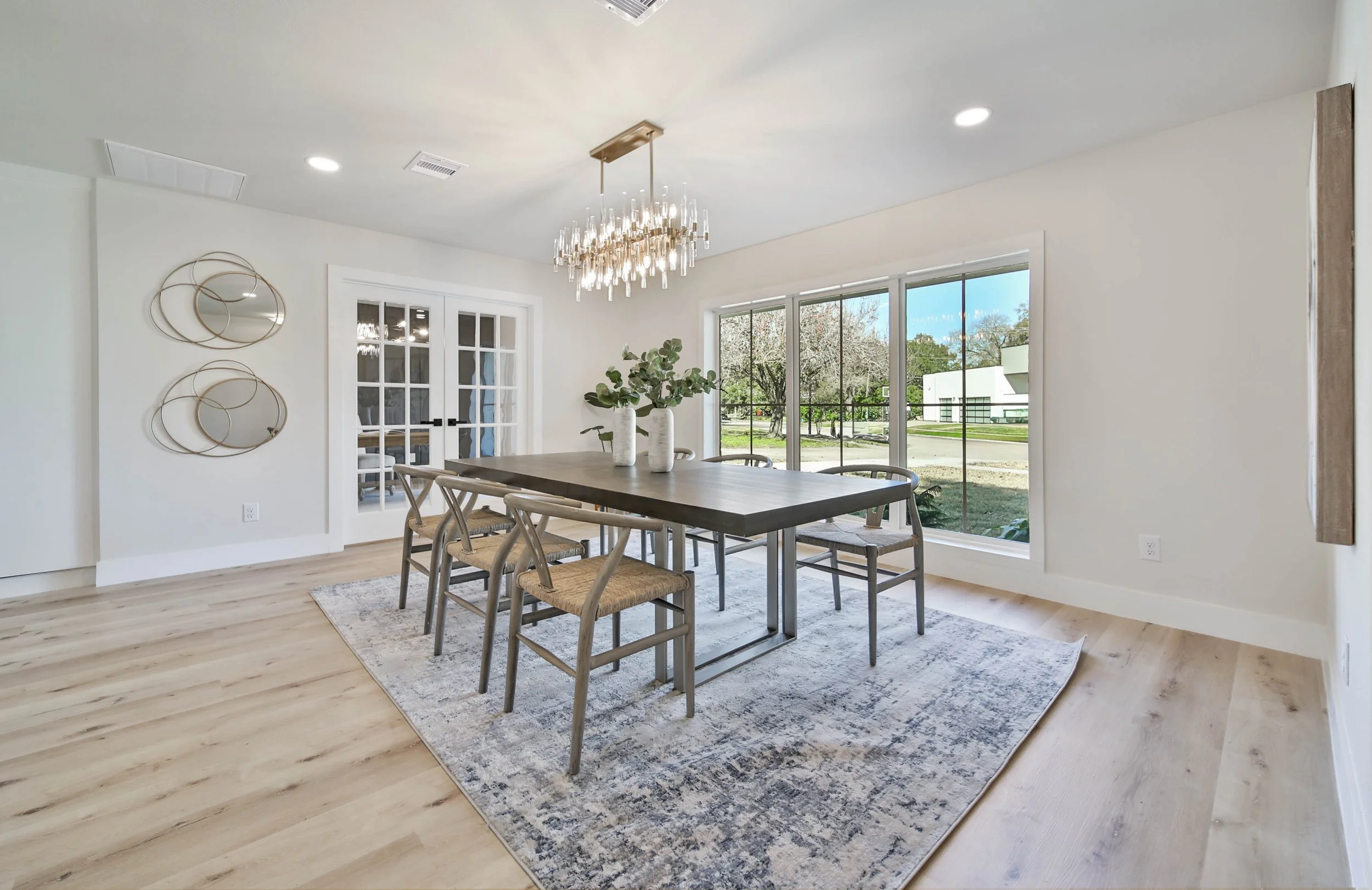 A modern dining room with a wooden table, six chairs, and a decorative pendant light fixture. Large windows offer a view of trees and a lawn outside, with a light-colored rug under the table. A wall features circular mirror art.