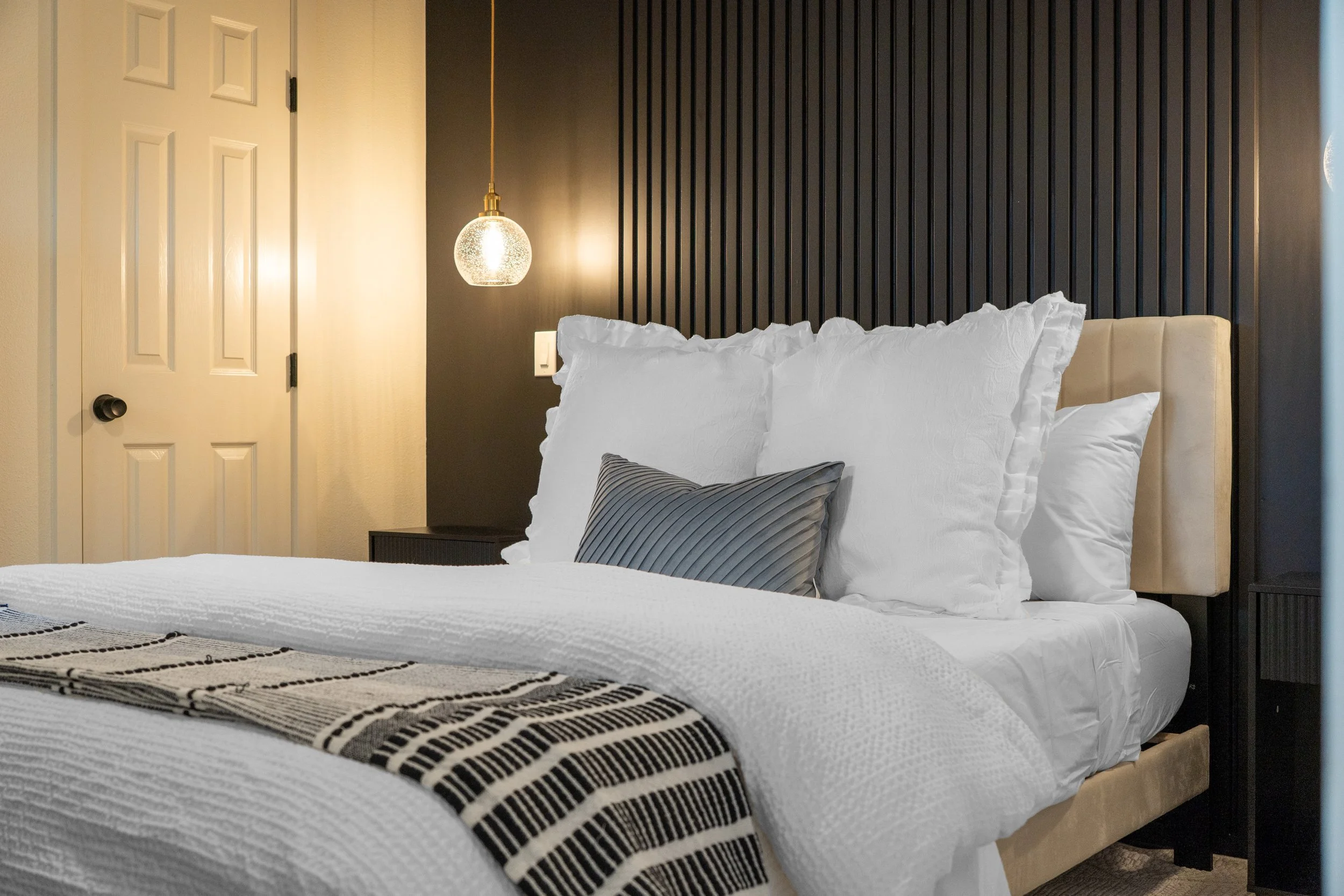 A bedroom with a bed, white bedding, and pillows. There is a black striped pillow, a bedside table, a hanging bedside lamp, and a black vertical slat wall behind the bed. A closed white door with black hardware is on the left.