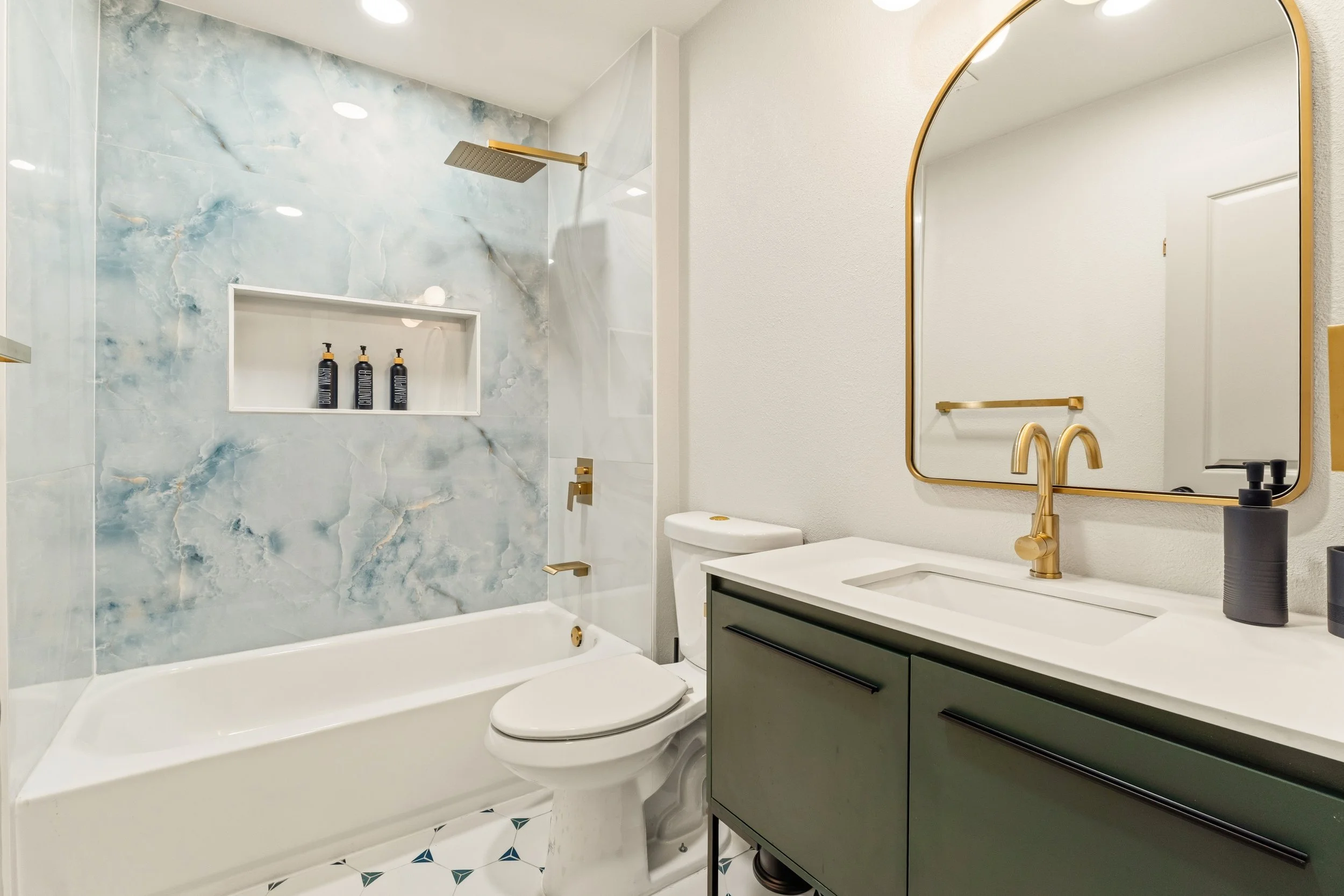 Modern bathroom with a bathtub and shower with blue marble tiles, a white toilet, a vanity with a sink and gold fixtures, a large mirror, and black soap dispensers.