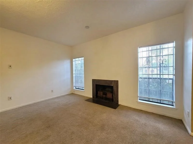 Empty living room with beige carpet, white walls, two windows with blinds, and a fireplace with a black mantel.
