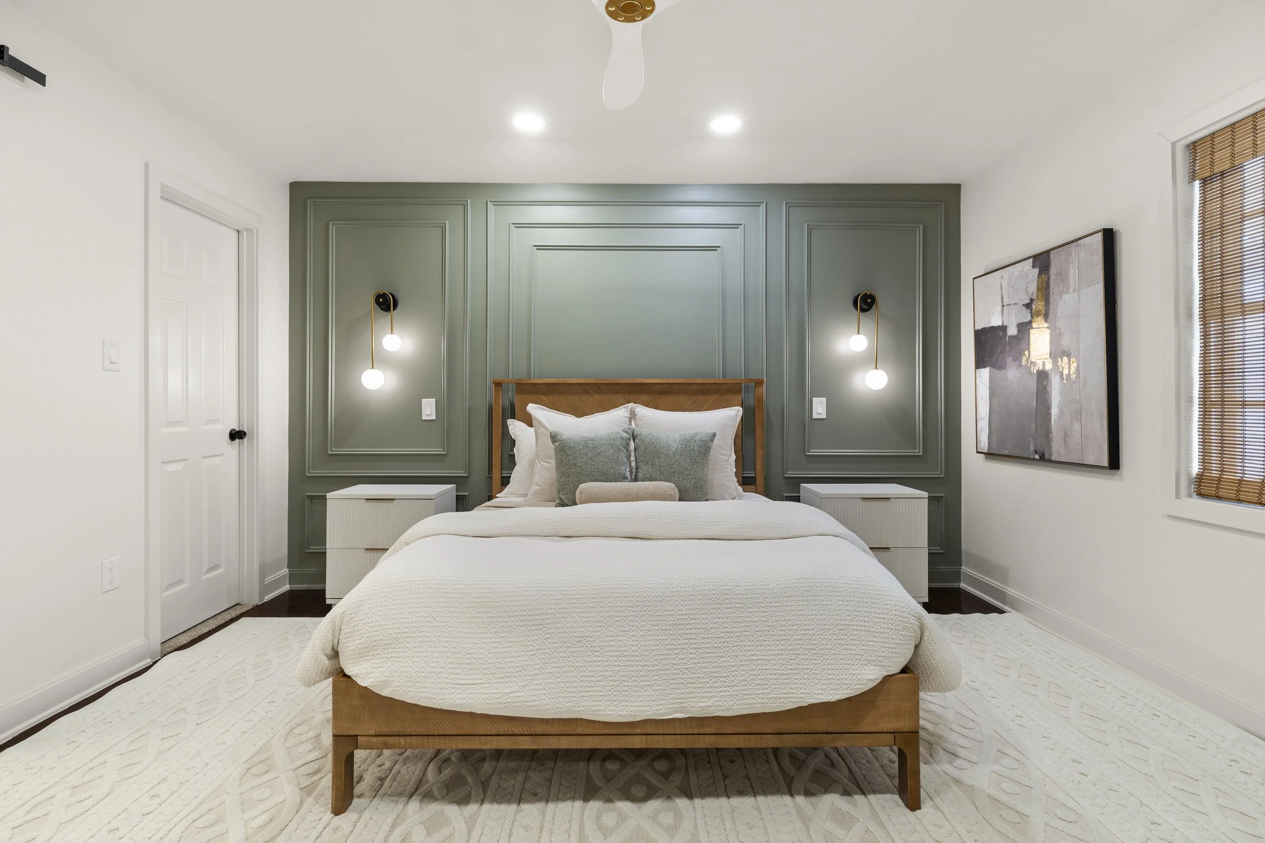 A modern bedroom featuring a bed with white bedding, green accent wall with molding, and symmetrical wall sconces. The room has white walls, a white door, and a window with bamboo blinds.