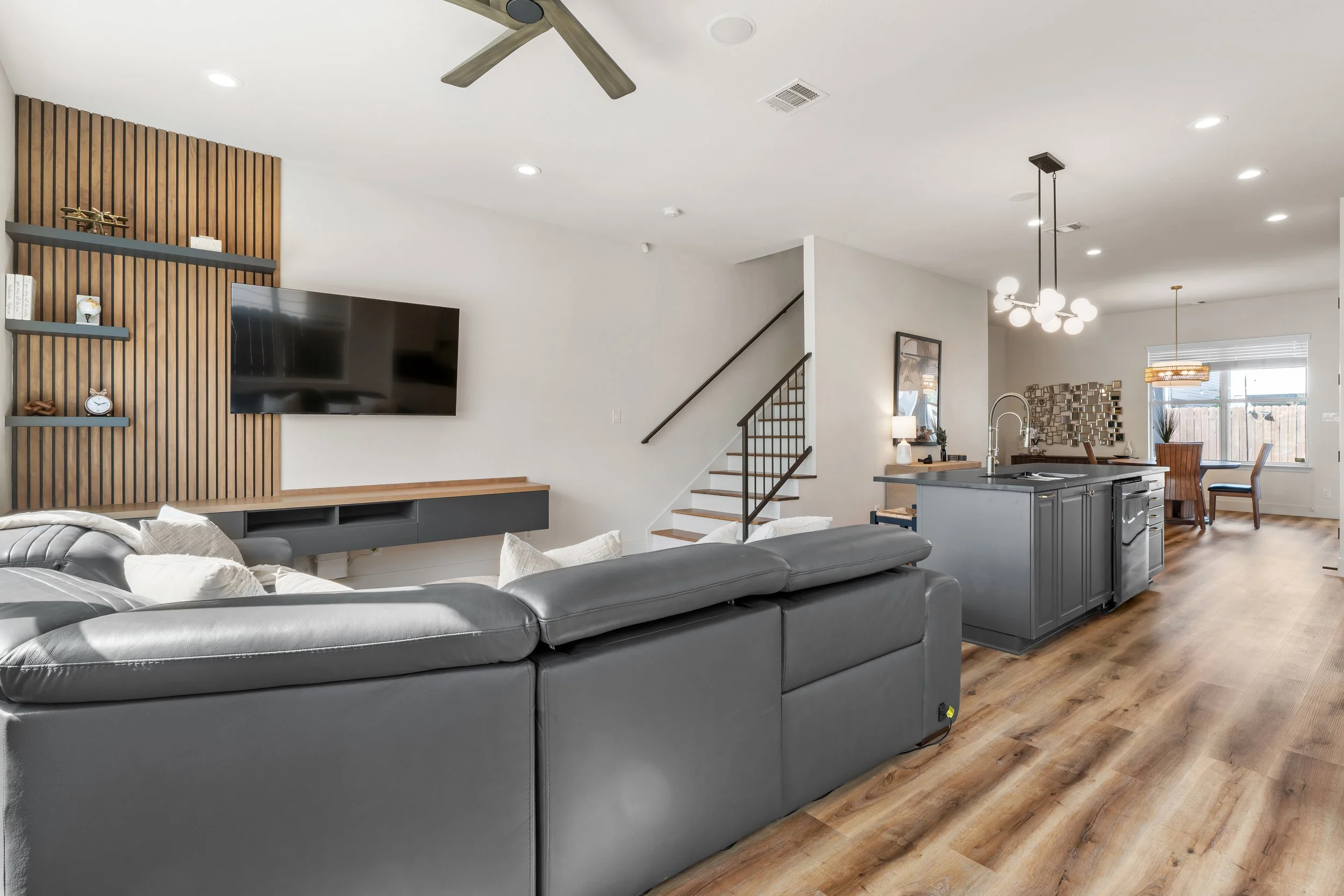 Open-concept living room and kitchen with gray leather sectional, TV mounted on wooden slat wall, staircase with black handrail, and dining area with pendant lights and window with blinds.