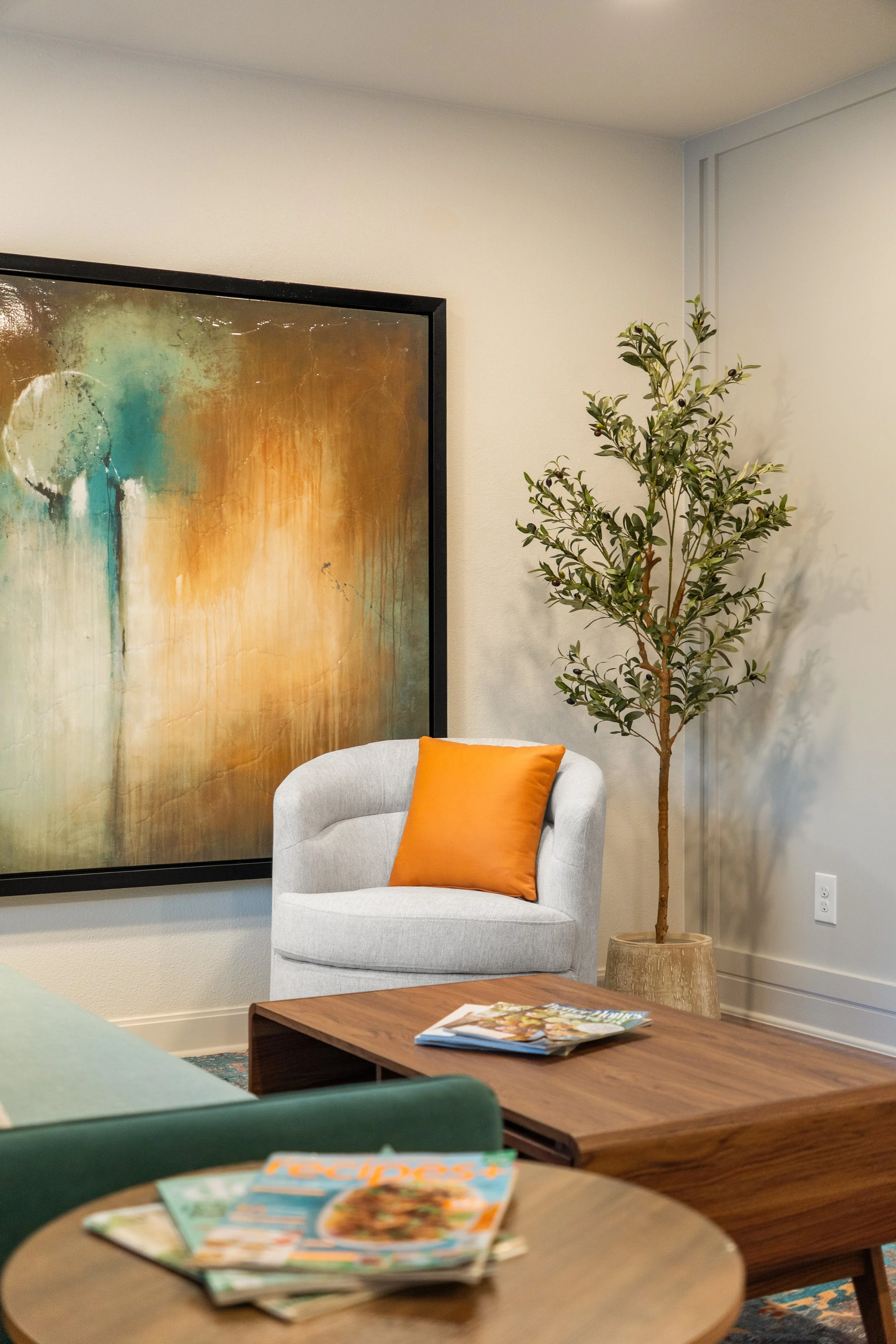 A cozy office or living room corner with a large abstract painting on the wall, a white armchair with an orange pillow, a tall potted plant, and wooden coffee tables with magazines on them.