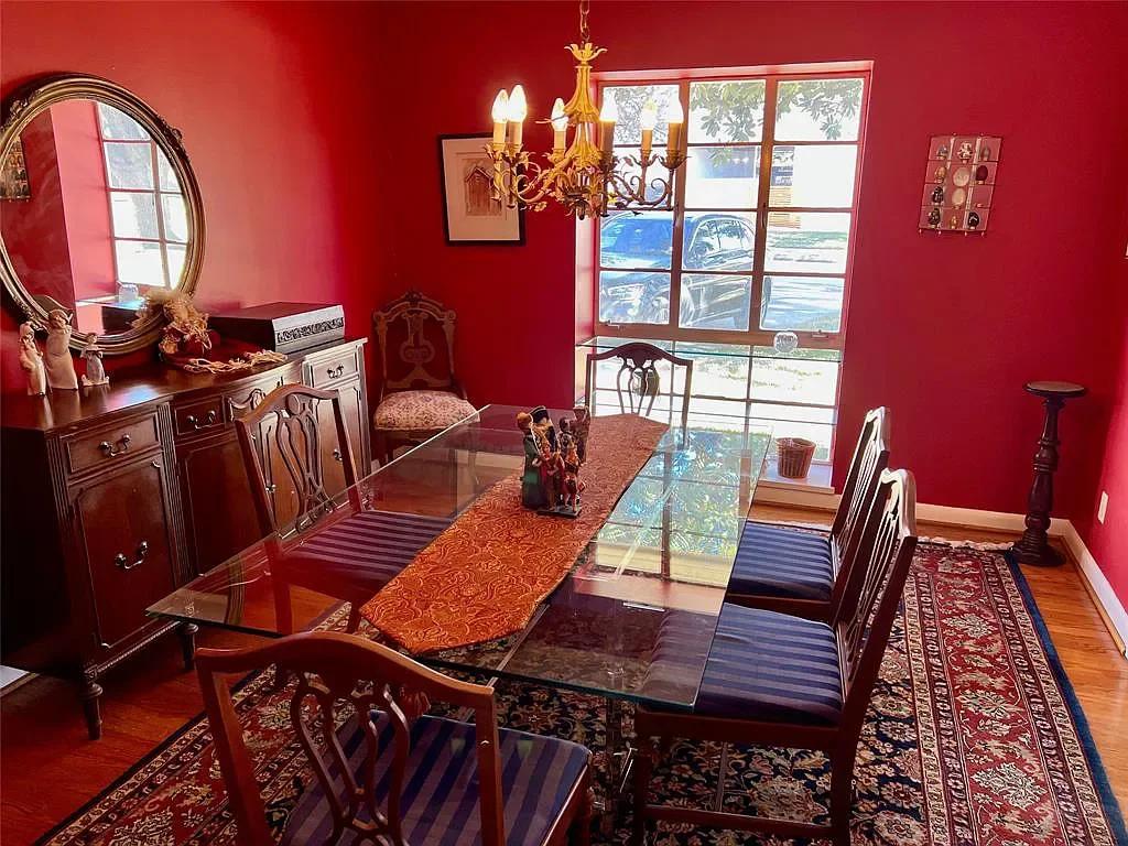 A dining room with red walls, a glass table, six wooden chairs, a wooden sideboard, a chandelier, a large window with sunlight, a patterned rug, and decorative items.